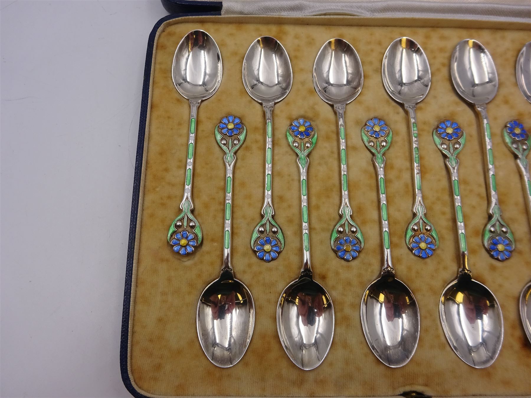 Set of twelve silver coffee spoons, each with enamelled blue flower to terminal and green and white enamel decoration to stem, hallmarked Mappin & Webb Ltd, Birmingham 1934, contained within fitted case