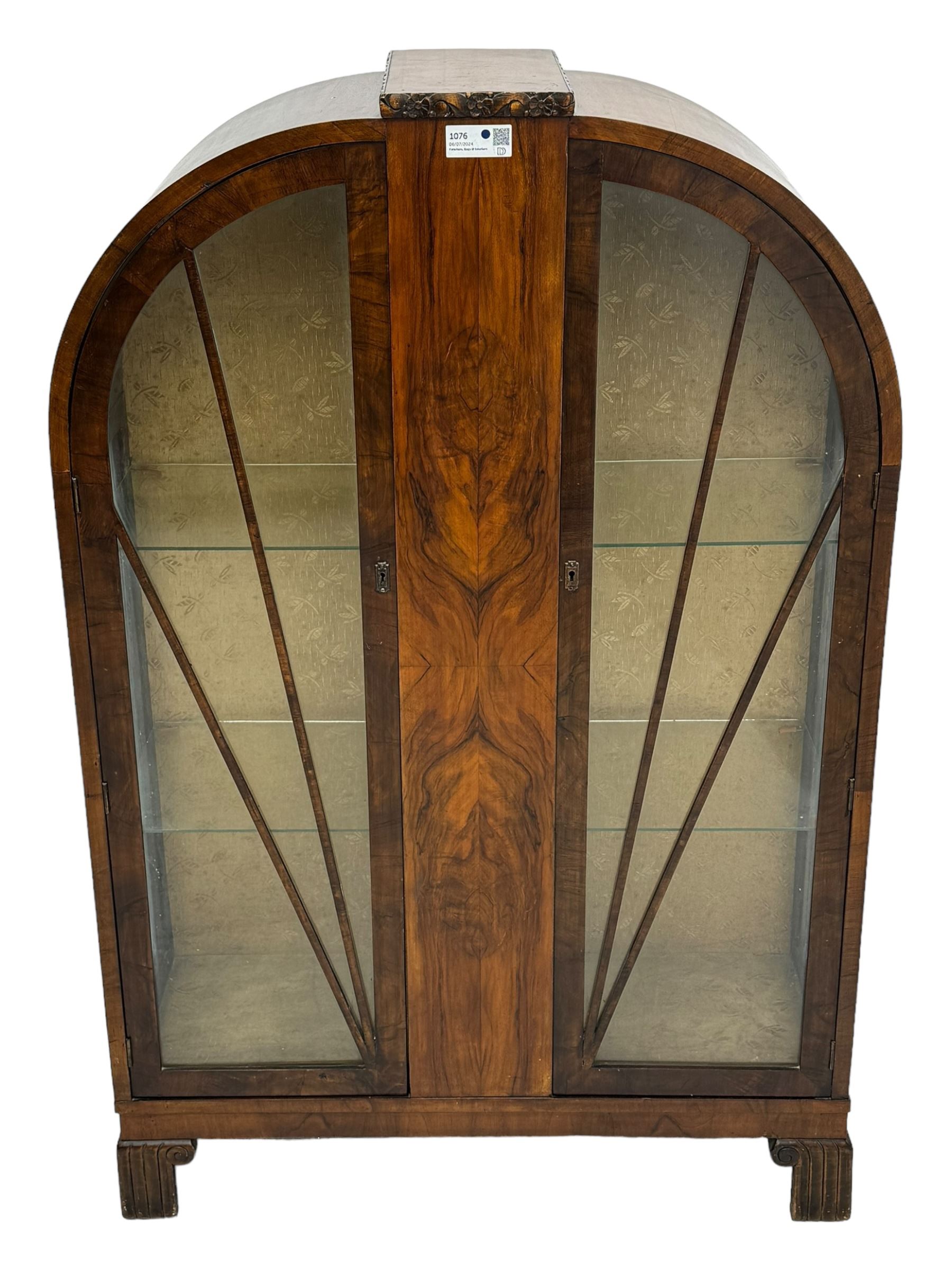 Early 20th century Art Deco walnut display cabinet, with arched top and carved floral crest, central band flanked by two glazed panel doors with decorative mullions, enclosing two glass shelves, on scroll carved bracket feet