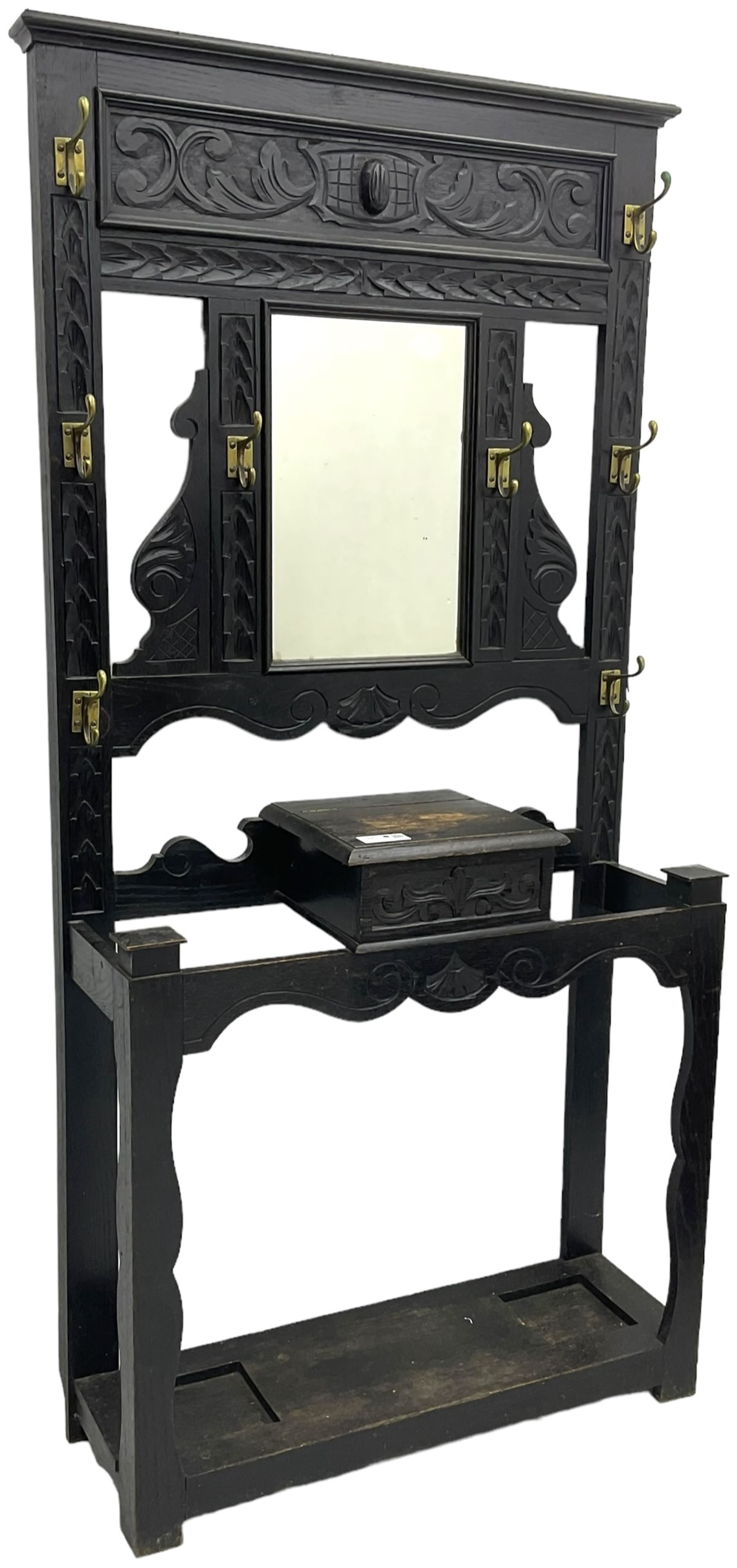 Victorian oak hall stand, the upper section featuring eight brass hooks and central mirror plate flanked by scroll-carved panels, the central section includes a storage box positioned above an umbrella stand, supported by square carved legs with scroll-carved horizontal supports