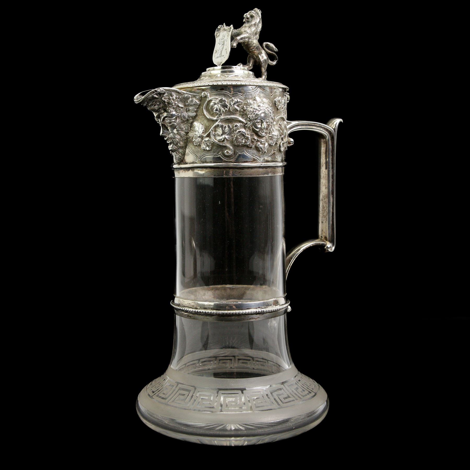 Victorian silver-mounted glass claret jug by George Richards Elkington, with Bacchanalian repoussé decoration and mask spout, with a domed hinged cover with a thumb-piece modelled as a rampant lion holding a shield, with engraved Dragon head crest, the glass body with etched greek key border and star-cut base, H29cm 