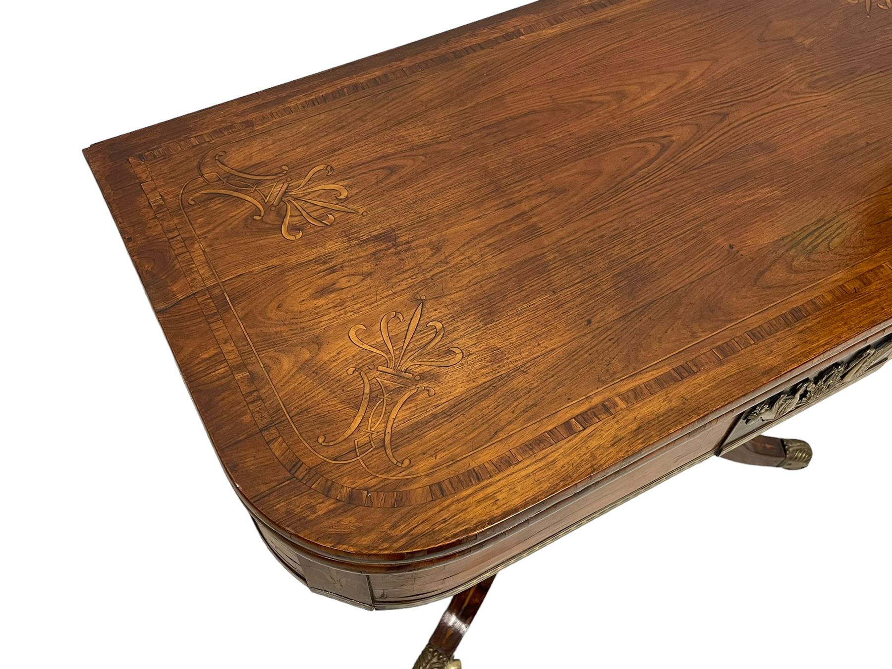 Regency inlaid rosewood card table, rectangular swivel and fold-over top with rounded corners and baize lined interior, the frieze decorated with a central ebony panel with applied gilt metal swan motifs, over a brass inlay band, raised on S-scroll supports united by concave platform base, terminating to out-splayed supports with brass cups and castors