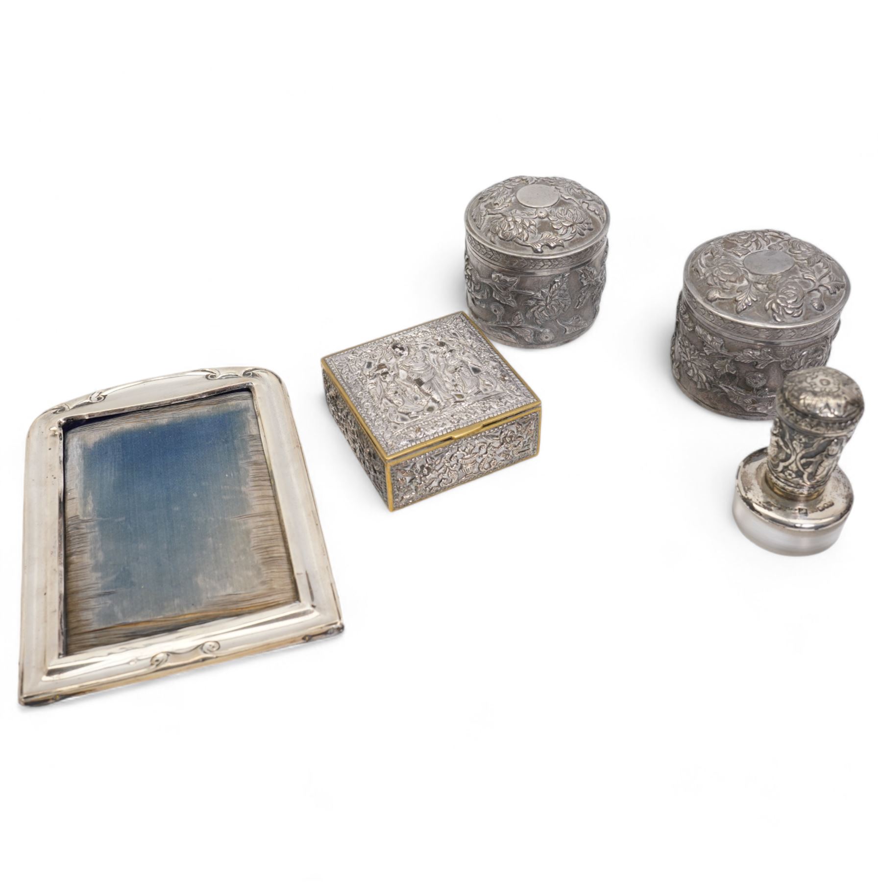 Pair of silver cylindrical dressing table boxes and covers embossed with trailing foliage H7cm, silver upright table photograph frame Birmingham 1909, Indian white metal and glass paperweight and a silver on copper trinket box