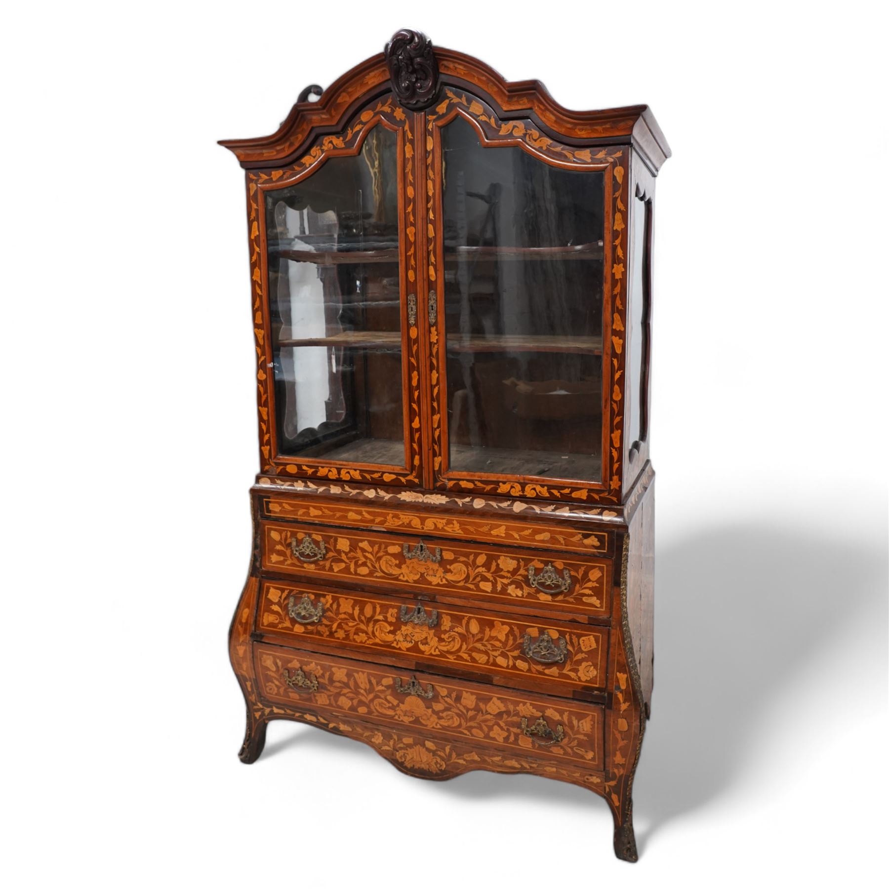 18th century Dutch marquetry walnut and gilt metal mounted vitrine cabinet on chest, shaped pediment carved with scrolling foliage cartouche, inlaid with trailing leafy branches and flower heads, two internal shelves enclosed by two glazed doors, the lower section with bombe outline fitted with three drawers, with ornate cast gilt metal edging and terminal caps