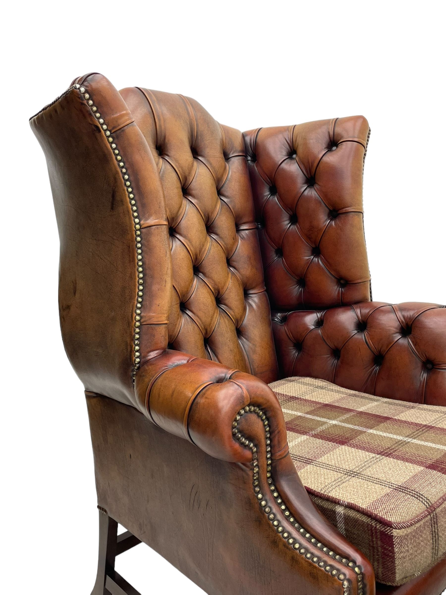 Georgian design hardwood-framed wingback armchair, shaped cresting rail and deep wingback over rolled arms, upholstered in brown buttoned leather with studwork bands, the seat cushion upholstered in checkered fabric, on square supports united by stretchers (W86cm, H118cm, D86cm); together with matching rectangular footstool (63cm x 43cm, H37cm) 