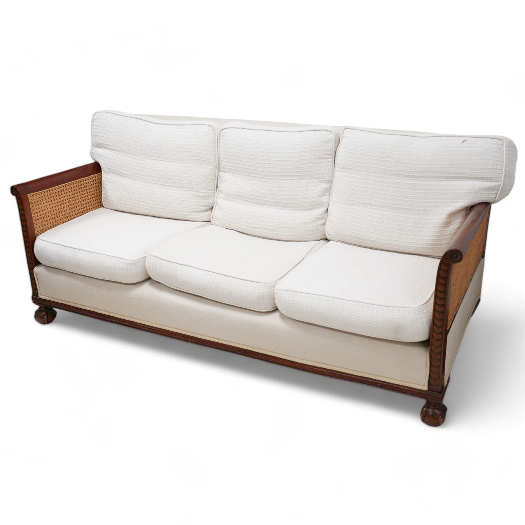 Early 20th century mahogany framed bergère three seat sofa, upholstered back and seat flanked by rolled double-caned arms with carved scale decoration, floral and ribbon-twist carved lower edge, over ball-and-claw feet