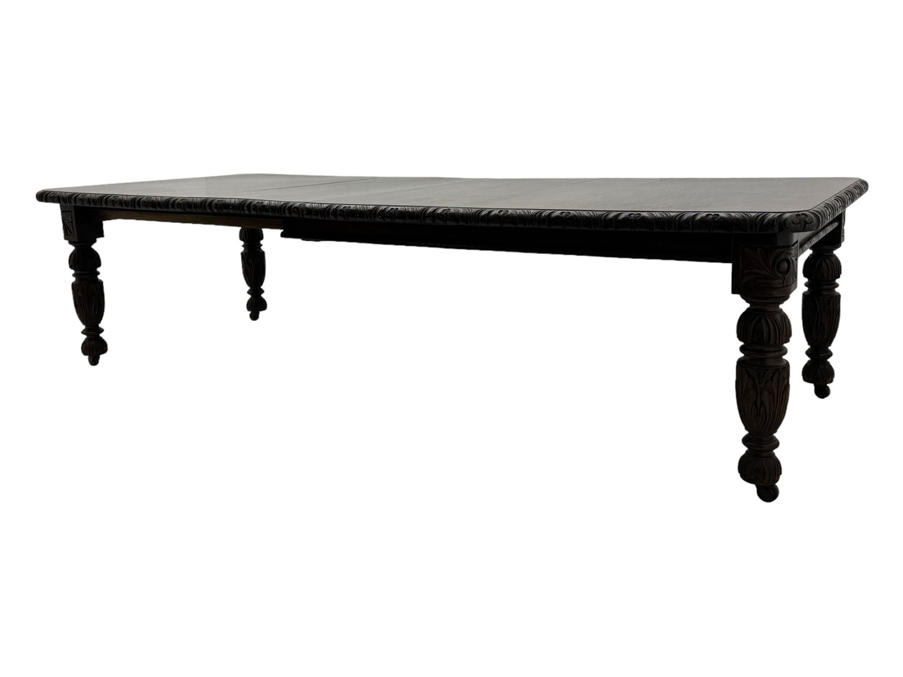 19th century heavily carved oak extending dining table, rectangular top with rounded corners and carved gadrooned edge, extending via winding mechanism with two additional leaves, raised on acanthus leaf-carved baluster supports terminating in ceramic castors