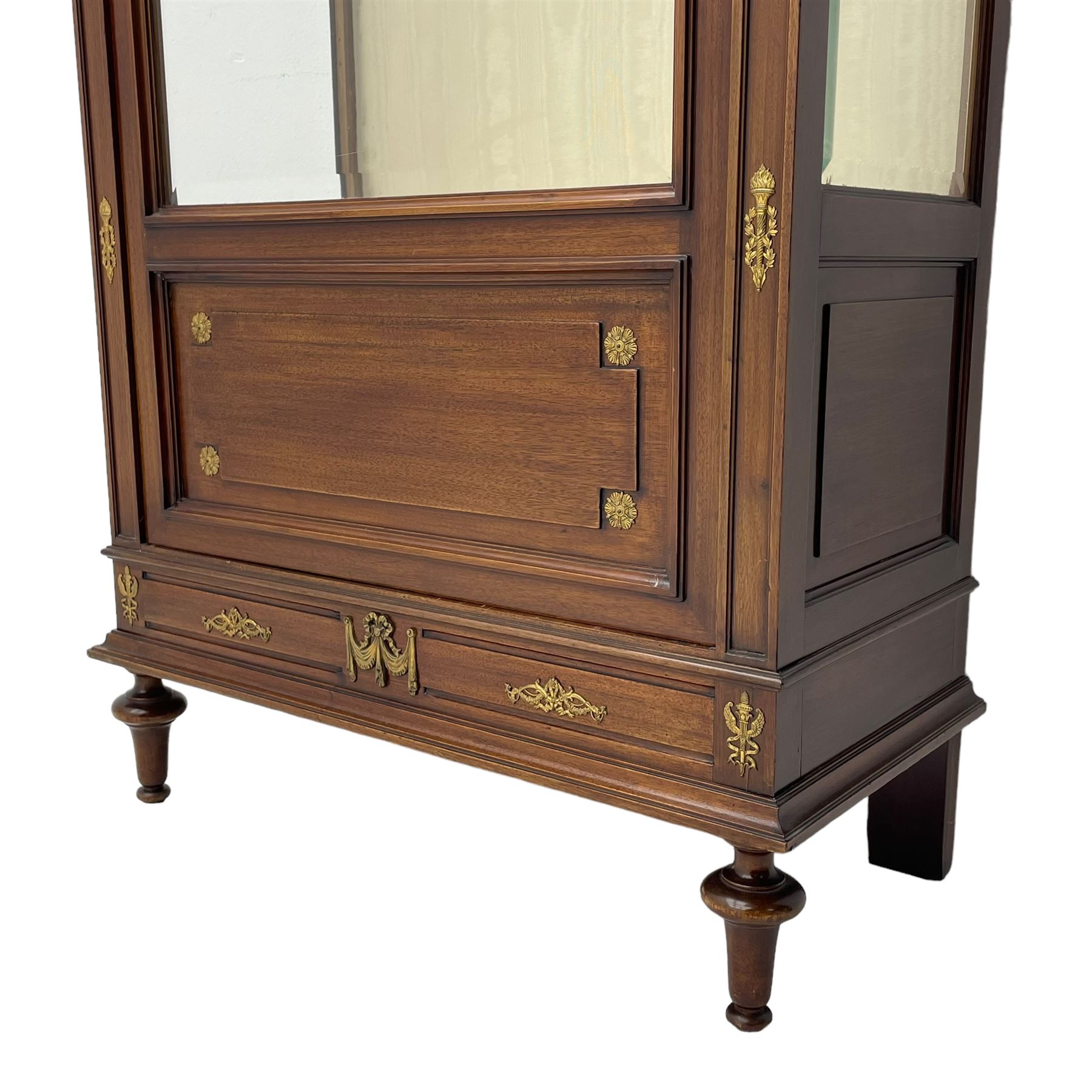 Early 20th century Louis XVI design mahogany vitrine display cabinet, projecting stepped cornice over recessed frieze decorated with gilt metal mounts of festoons and a bellflower wreath, fabric lined interior with two glass shelves, enclosed by bevel glazed door with lower panel, on two turned front feet 