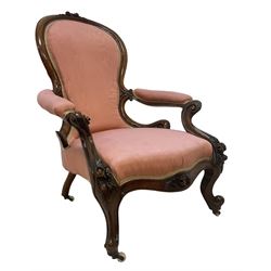 Victorian mahogany framed open armchair, spoon-back and sprung seat upholstered in pink fabric, scrolled arm terminals over serpentine fronted seat decorated with moulded carved floral motifs, cabriole supports with ceramic castors