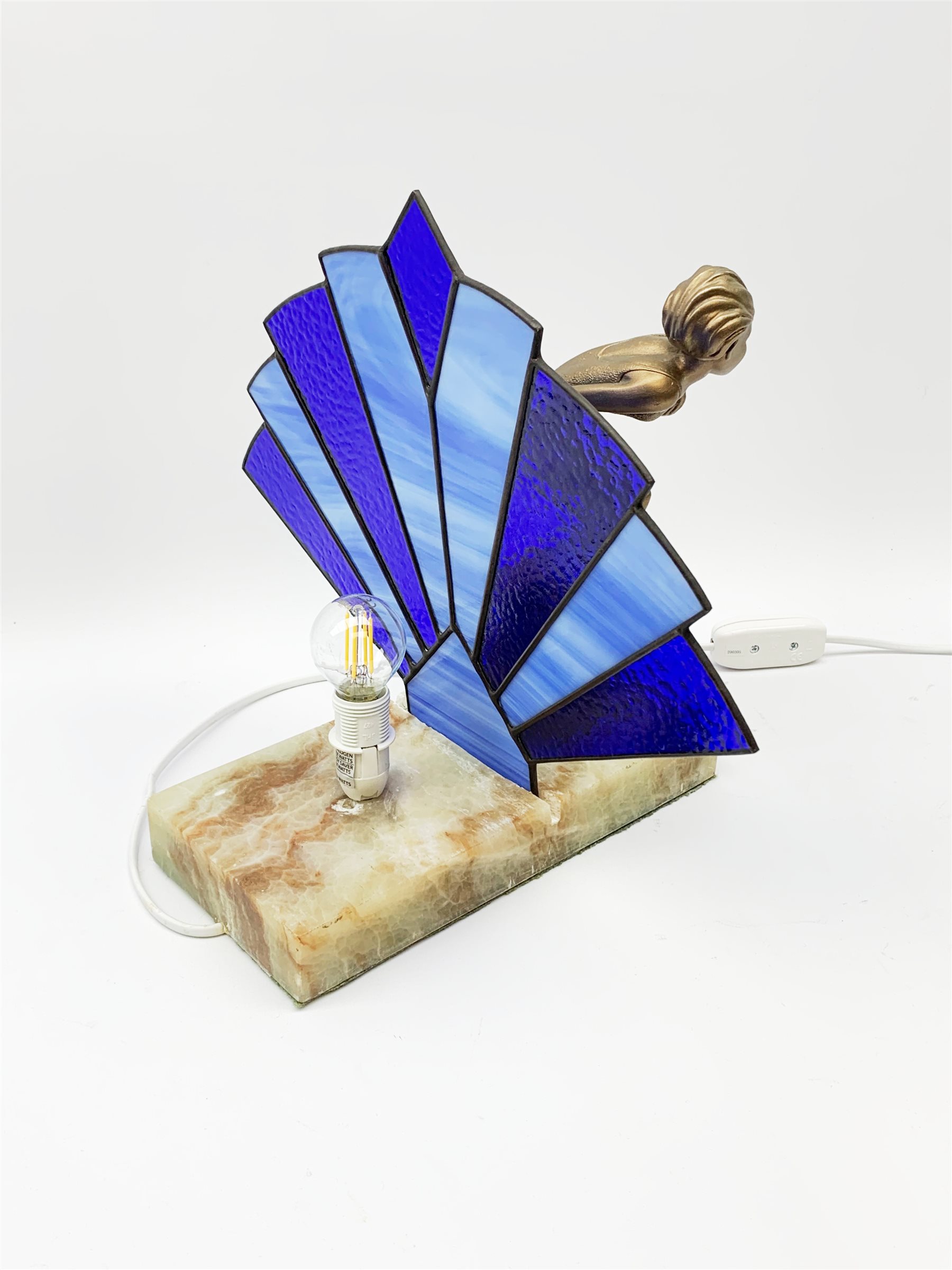 An Art Deco style table lamp, modelled in the form of a bronzed female figure before a leaded blue glass fan shaped shade, upon rectangular onyx base, overall H32cm base L28cm. 