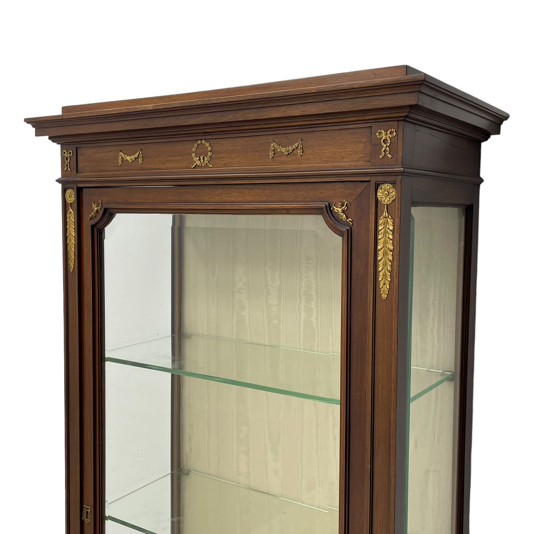 Early 20th century Louis XVI design mahogany vitrine display cabinet, projecting stepped cornice over recessed frieze decorated with gilt metal mounts of festoons and a bellflower wreath, fabric lined interior with two glass shelves, enclosed by bevel glazed door with lower panel, on two turned front feet 