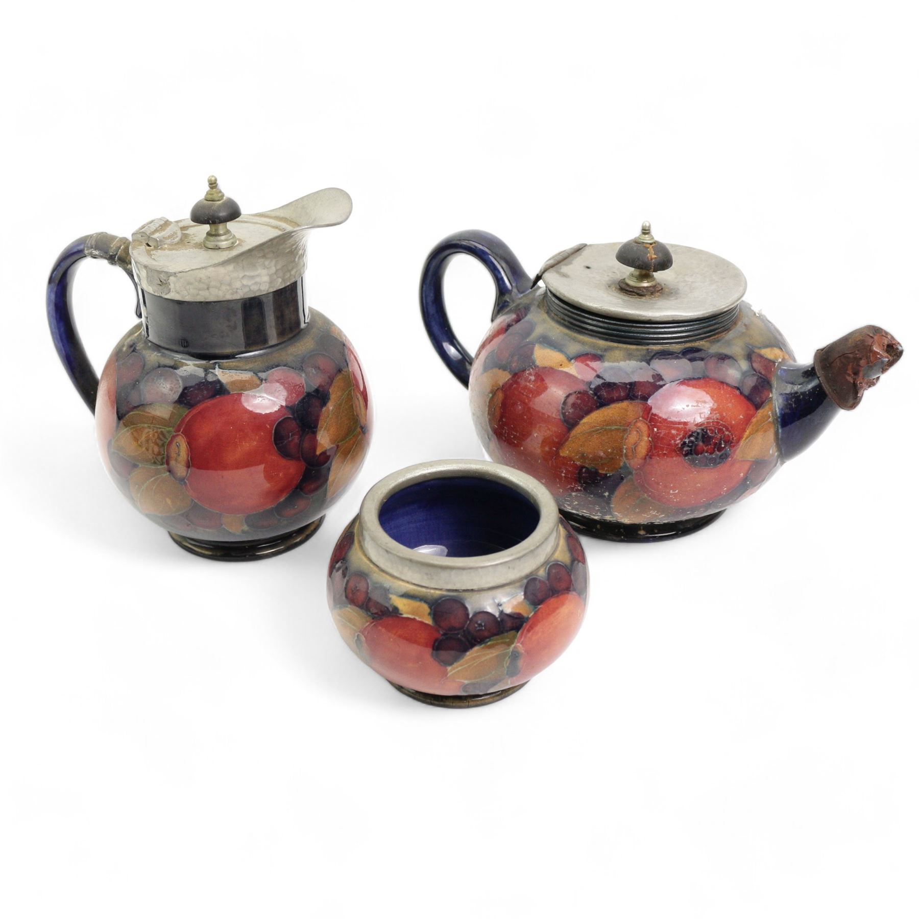 William Moorcroft Pomegranate pattern three piece tea service, comprising teapot with hammered pewter mounts and lid, sugar basin and hot water pot, all with pewter hammered finish mounts, possibly designed for Liberty & Co. 