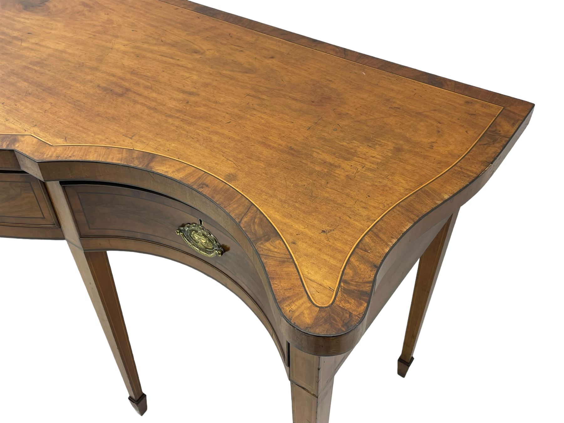 Large George III mahogany serpentine serving table, shaped top with crossing banding and stringing, the frieze fitted with three cock-beaded drawers, oval pressed brass handle plates decorated with urns, on square tapering supports with spade feet 