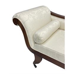 Regency design mahogany chaise longue, scrolled arms with carved rosette details, upholstered in cream damask fabric with bolster cushion, reeded frame supported by turned legs on brass castors