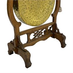 Early 20th century oak gong stand with gong and hammer, shaped cresting rail over moulded upper edge on shaped supports, hanging brass drum gong over fretwork middle rail, on raised platforms terminating to splayed feet