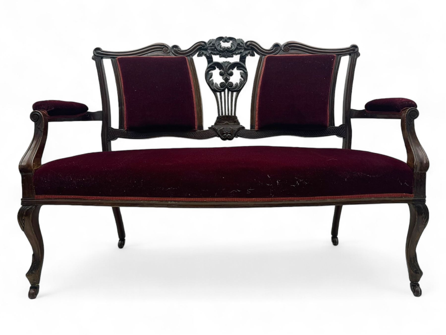 Late Victorian dark oak framed sofa, the backrests, seat and padded arms upholstered in deep red fabric, carved crest rail with scroll and foliate motifs supported by carved uprights, on cabriole supports with leaf carvings terminating in castors