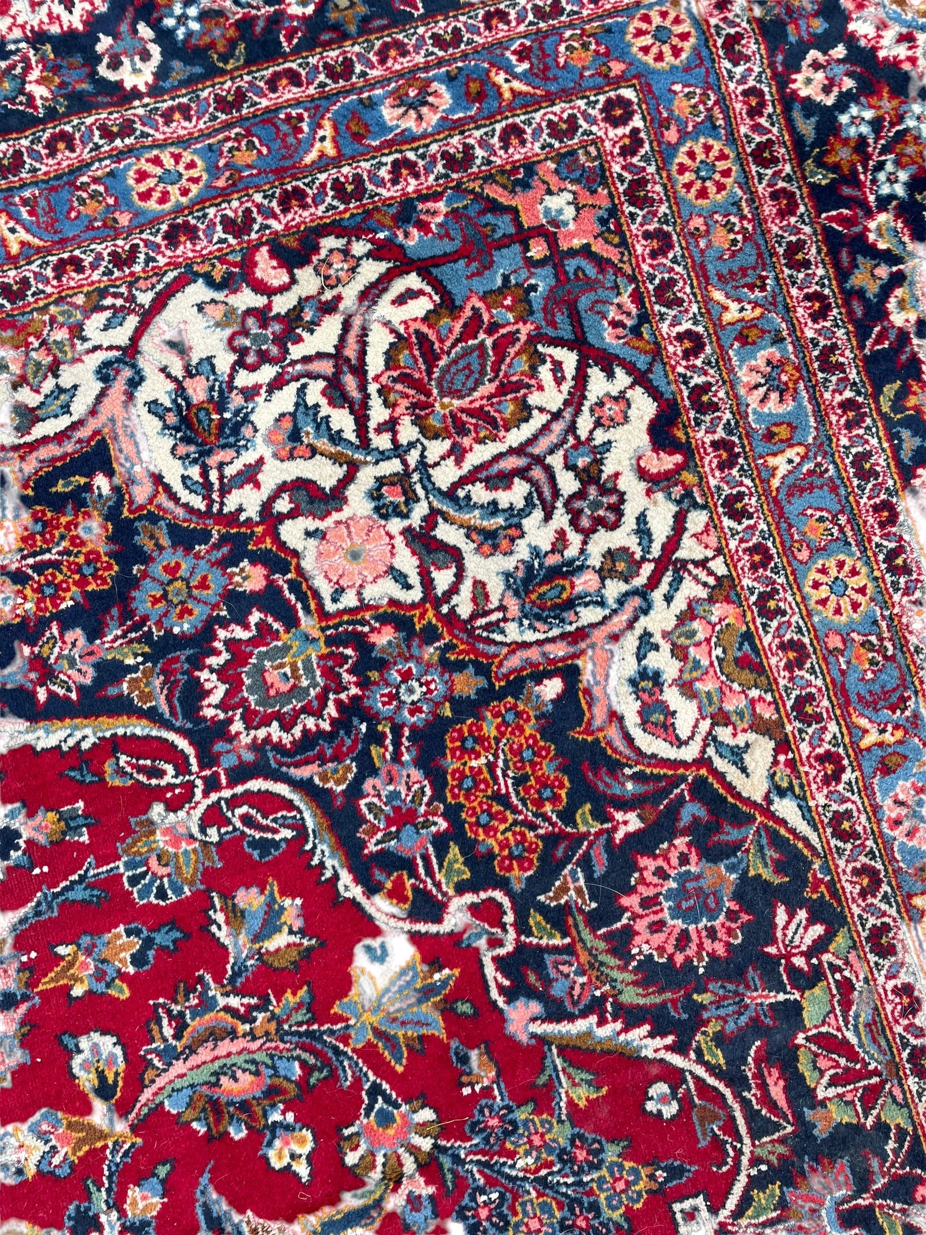 Large Persian Kashan crimson ground carpet, central floral medallion surrounded by swirling leafy branches and palmettes, enclose by floral pattern spandrels, the indigo border with overall scrolling design decorated with palmettes, within guard stripes 