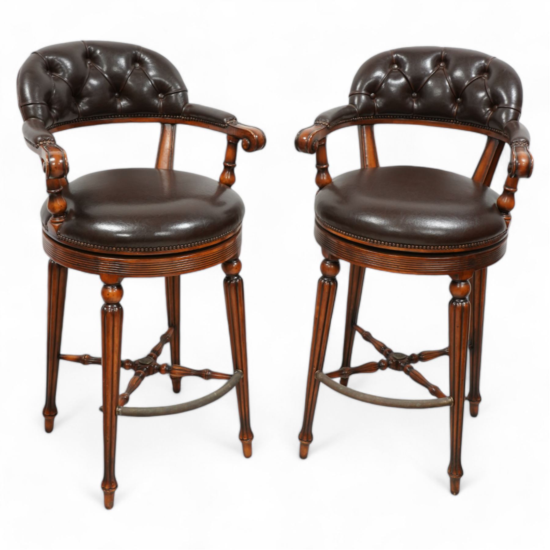 Pair of hardwood-framed bar stools, revolving tub seat with buttoned back rest and upholstered seat in brown leather, reed moulded seat rail over four lobe carved tapering supports, united by metal foot rest and x-framed stretchers 