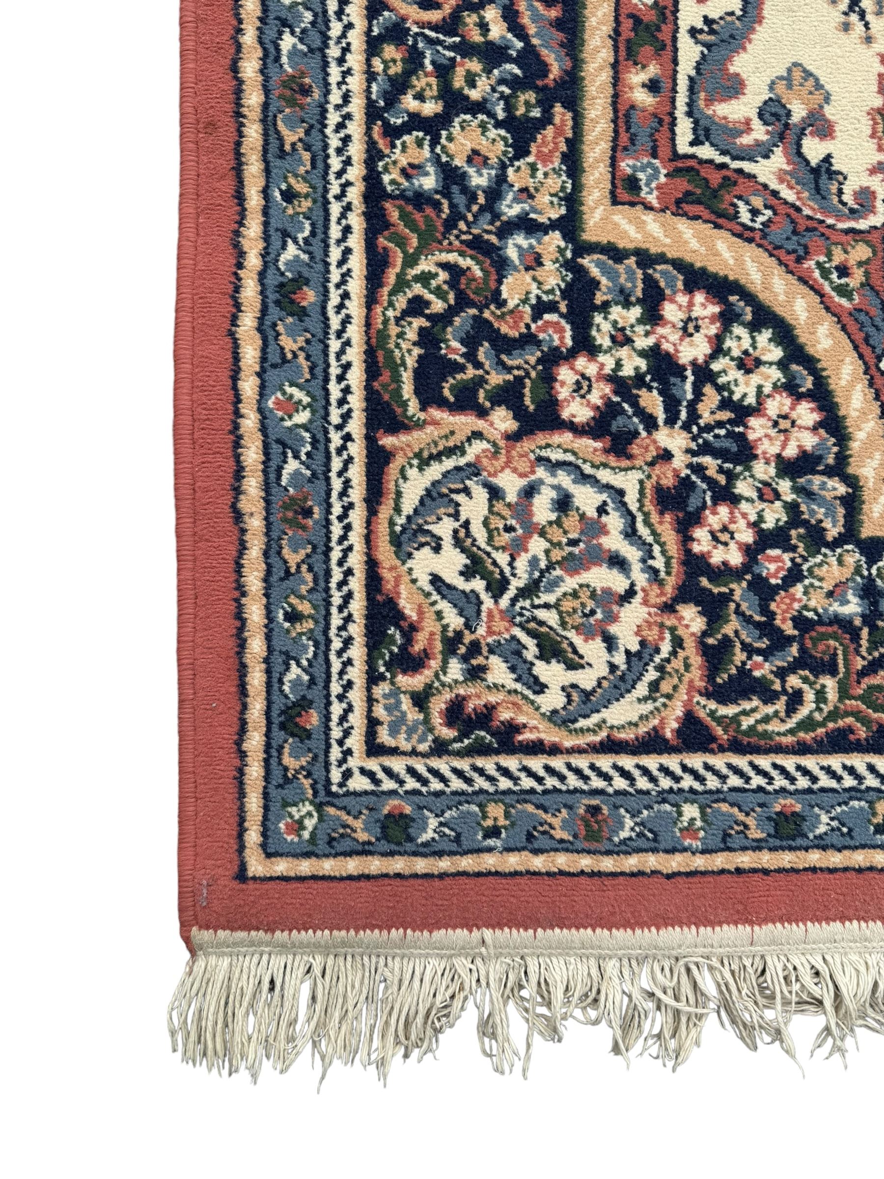 Persian design ivory ground rug, the field decorated with an oval medallion filled with floral motifs and scrolling vines, flanked by floral sprays and corner spandrels with palmette designs, the main border featuring a series of stylised floral patterns against a dark blue ground, enclosed by multiple guard stripes with geometric and floral motifs