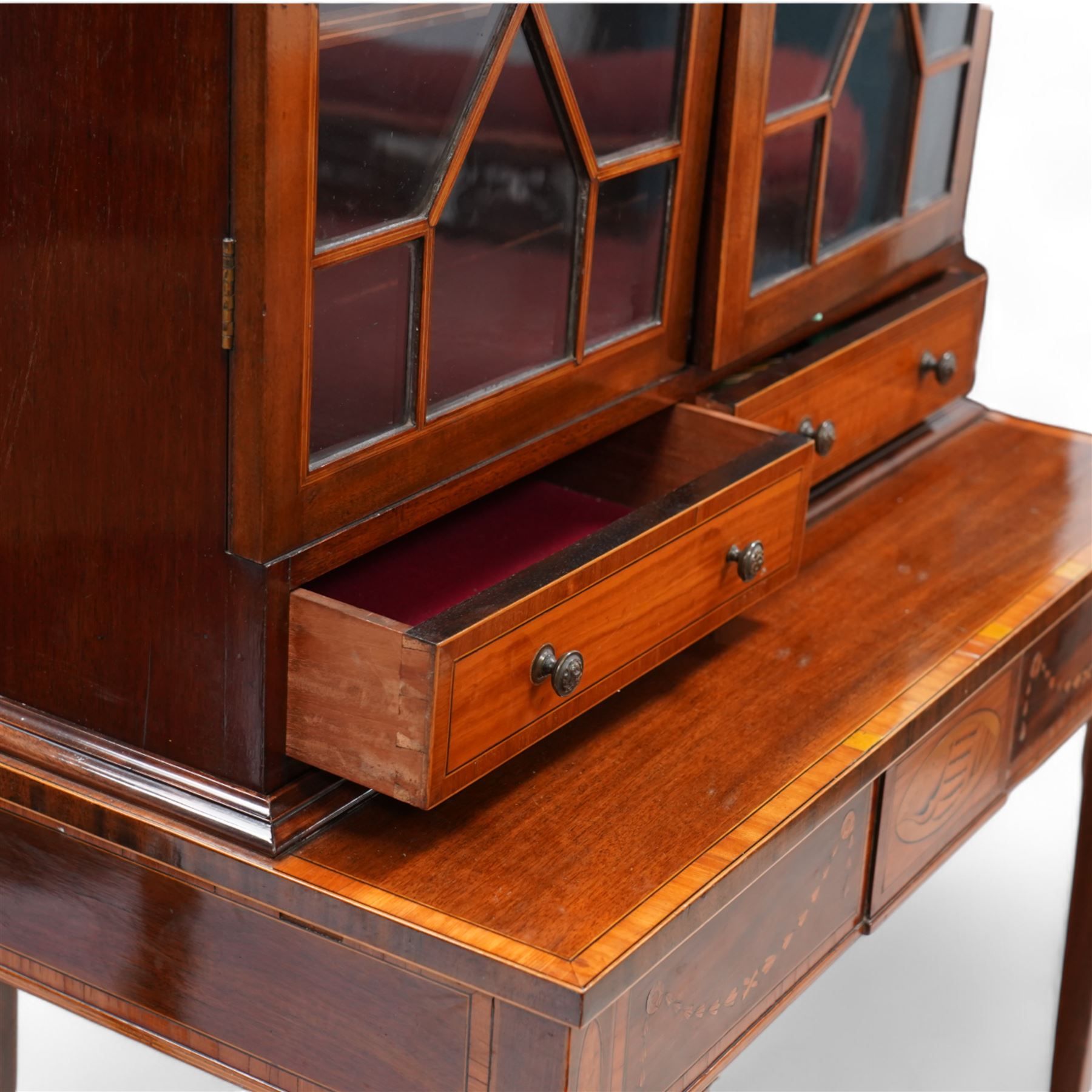 Early 19th century mahogany and satinwood bookcase on stand, scrolled swan neck pediment with pierced panels, satinwood frieze over two astragal glazed doors enclosing adjustable shelves, fitted with two drawers, the stand with rectangular satinwood banded top over frieze inlaid with central shell panel and extending bell flower festoons, on square tapering supports united by undertier with raised gallery 