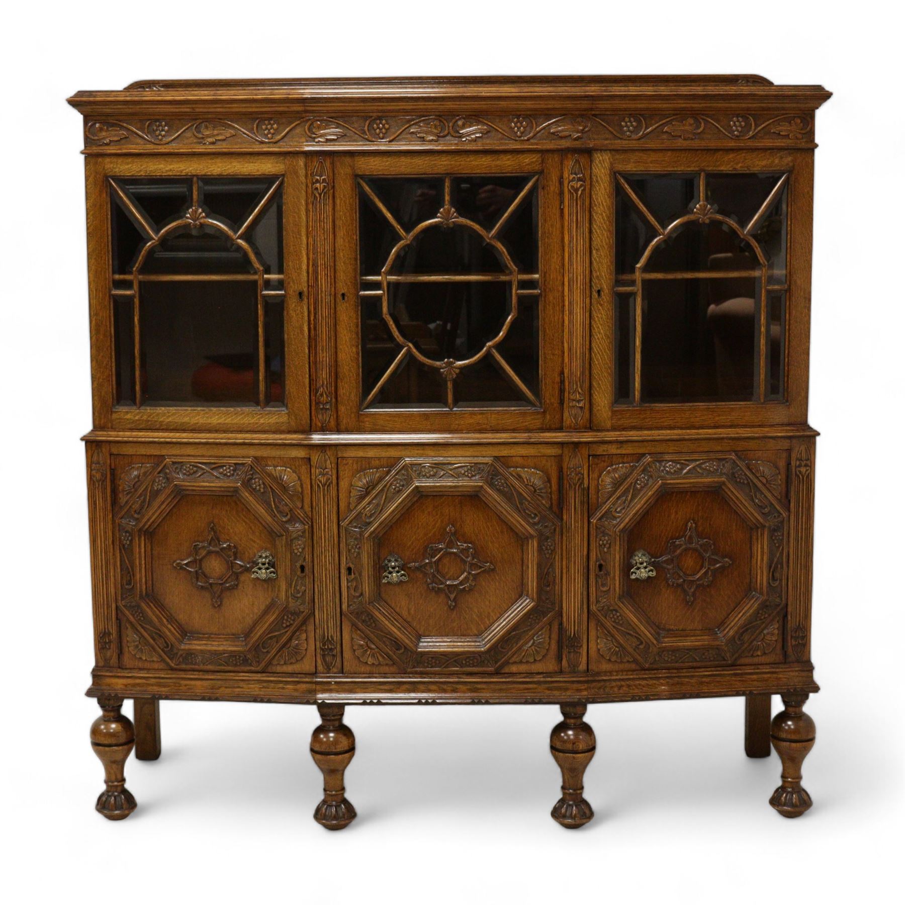 Early 20th century heavily carved oak display cabinet, frieze carved with grape vines over three astragal glazed doors with bevelled glass, over three cupboards applied with octagonal carved panels with further vine carvings, on turned and lobe carved supports