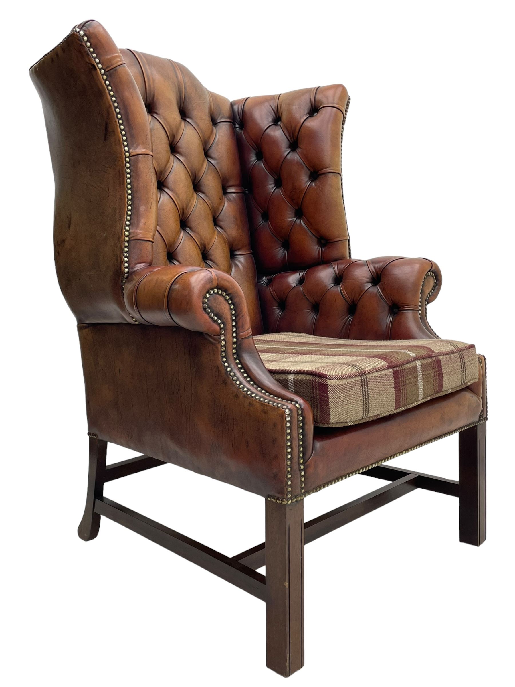 Georgian design hardwood-framed wingback armchair, shaped cresting rail and deep wingback over rolled arms, upholstered in brown buttoned leather with studwork bands, the seat cushion upholstered in checkered fabric, on square supports united by stretchers (W86cm, H118cm, D86cm); together with matching rectangular footstool (63cm x 43cm, H37cm) 