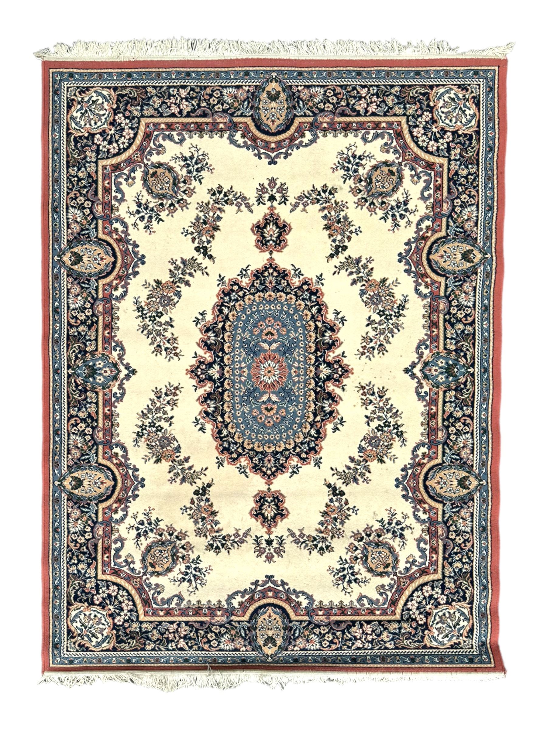 Persian design ivory ground rug, the field decorated with an oval medallion filled with floral motifs and scrolling vines, flanked by floral sprays and corner spandrels with palmette designs, the main border featuring a series of stylised floral patterns against a dark blue ground, enclosed by multiple guard stripes with geometric and floral motifs