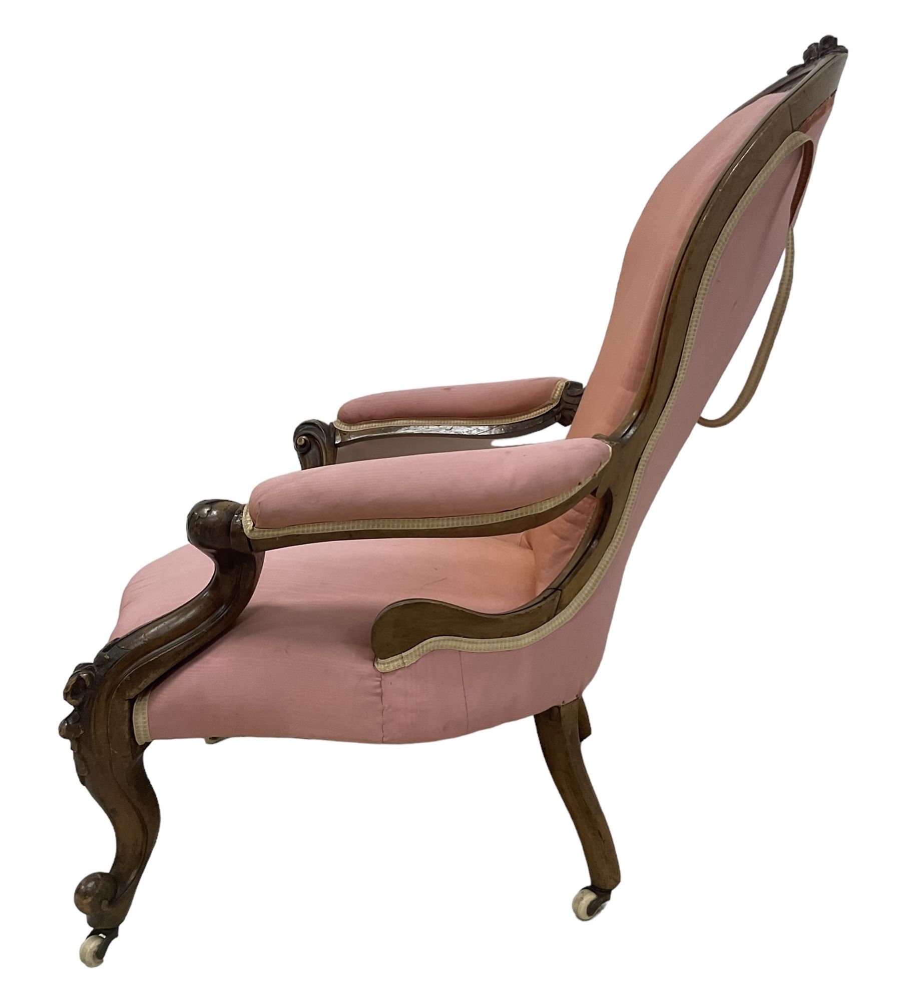 Victorian mahogany framed open armchair, spoon-back and sprung seat upholstered in pink fabric, scrolled arm terminals over serpentine fronted seat decorated with moulded carved floral motifs, cabriole supports with ceramic castors