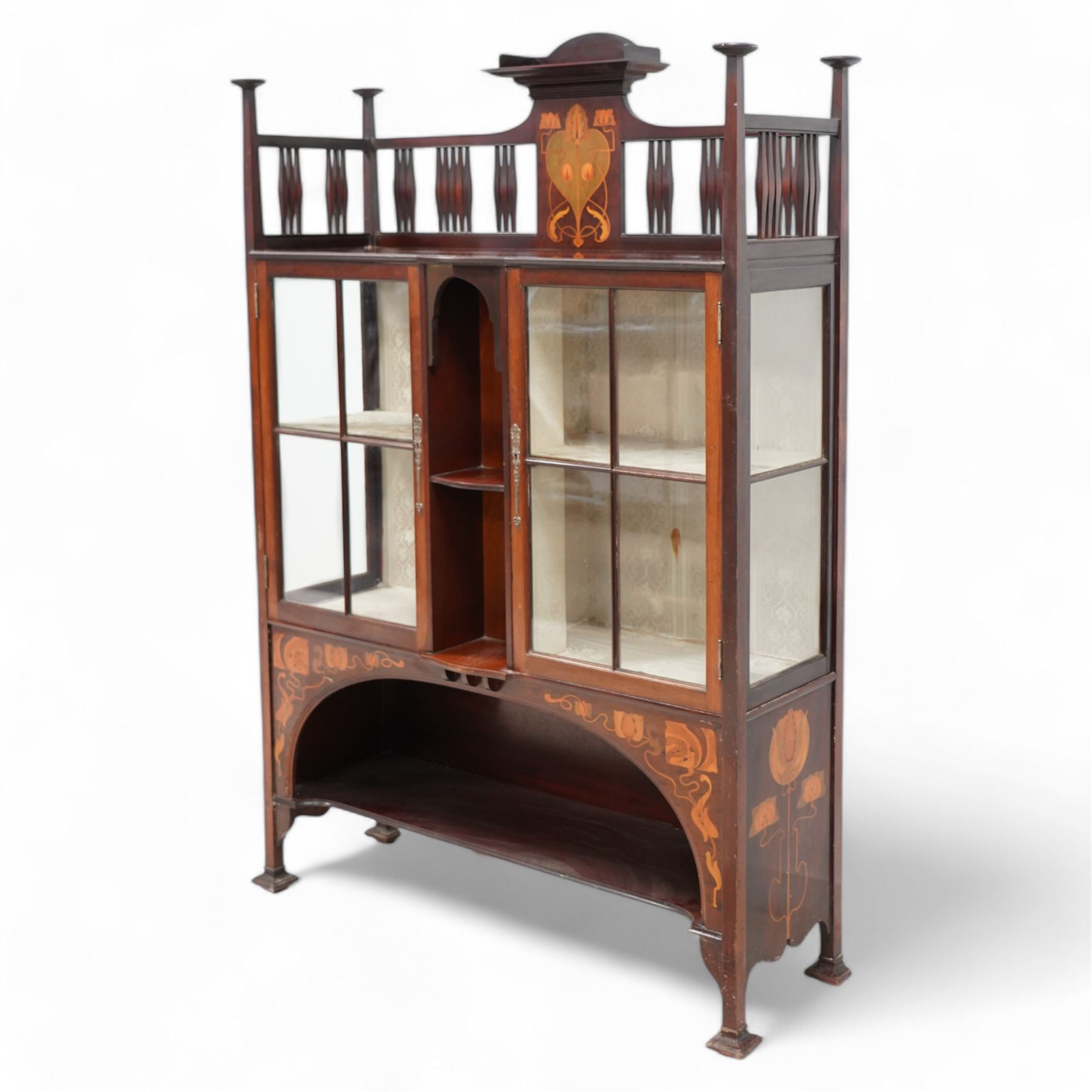 Shapland & Petter; Barnstaple (1854-1999) - Art Nouveau circa. 1900s mahogany display cabinet, raised shelf over panel inlaid with heart and stylised tulips with curled leaves, flanked by balustrade, enclosed by two astragal glazed doors, the lined interior fitted with two shelves, recessed central upright fitted with single shelf, arched lower section fitted with undertier, the spandrels inlaid with further stylised flower heads and trailing branches, on style supports terminating to moulded pad feet, the locks stamped 'S&B, B', retailed by 'Denby & Spinks, Leeds' plaque verso