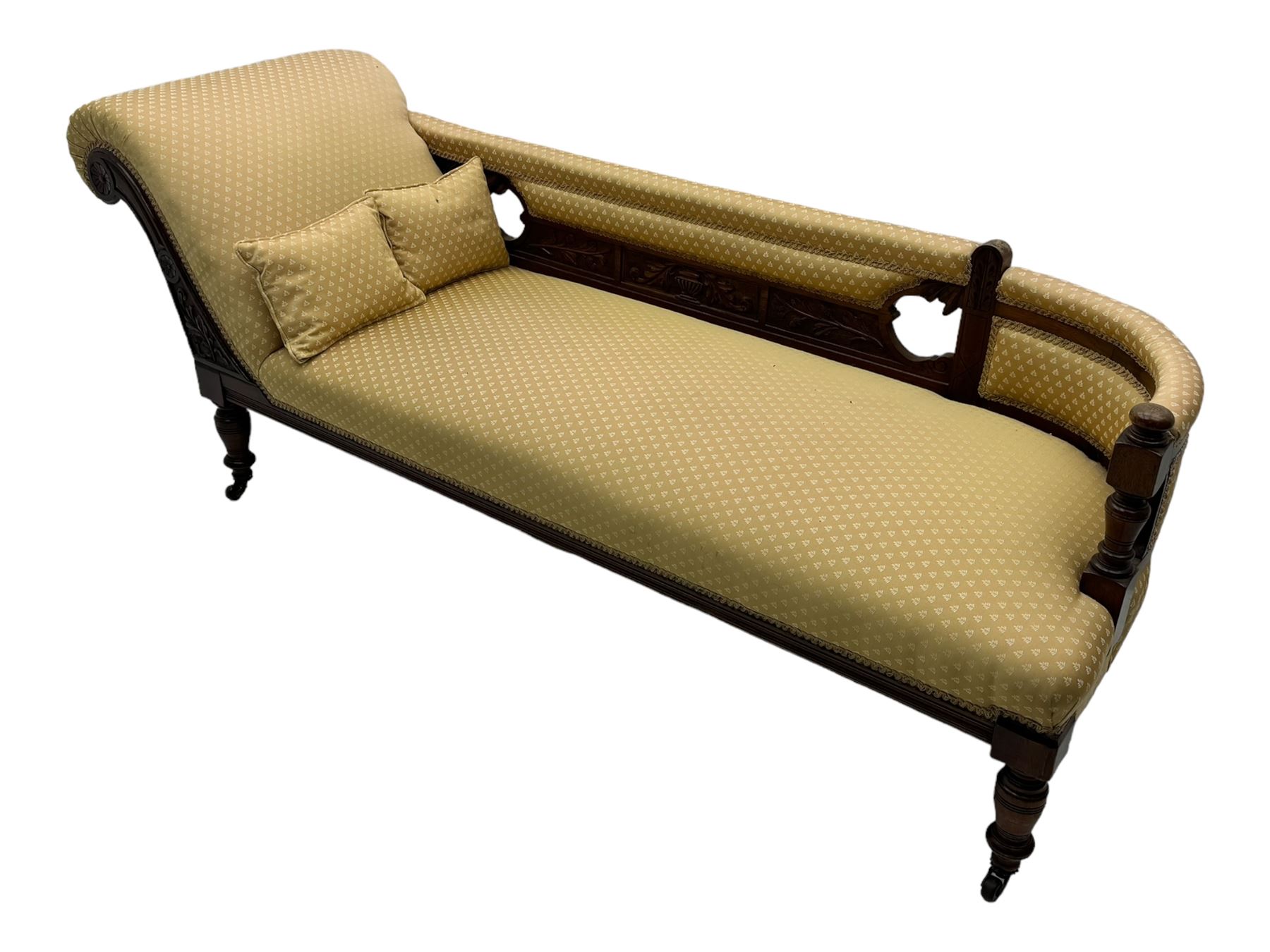 Late Victorian walnut framed chaise longue, scrolled backrest and sides upholstered in patterned yellow fabric, carved floral motifs to the backrest and sides, raised on turned supports with castors