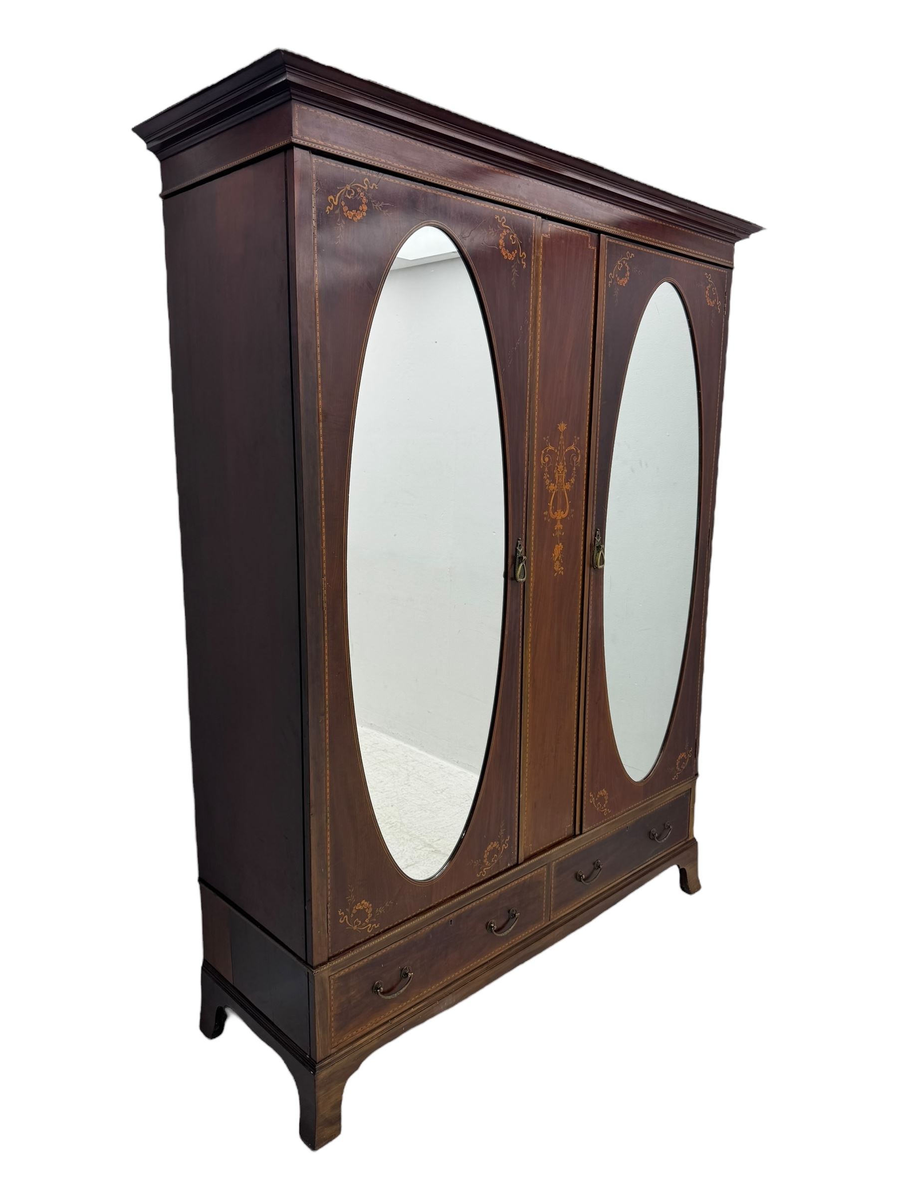 Maples & Co. - Edwardian mahogany wardrobe, projecting cornice over frieze inlaid with checkered stringing, central vertical panel inlaid with lyre and scrolling foliate decoration, two oval mirror glazed doors enclosing hanging rails and hooks, the base fitted with two short drawers, on splayed bracket feet