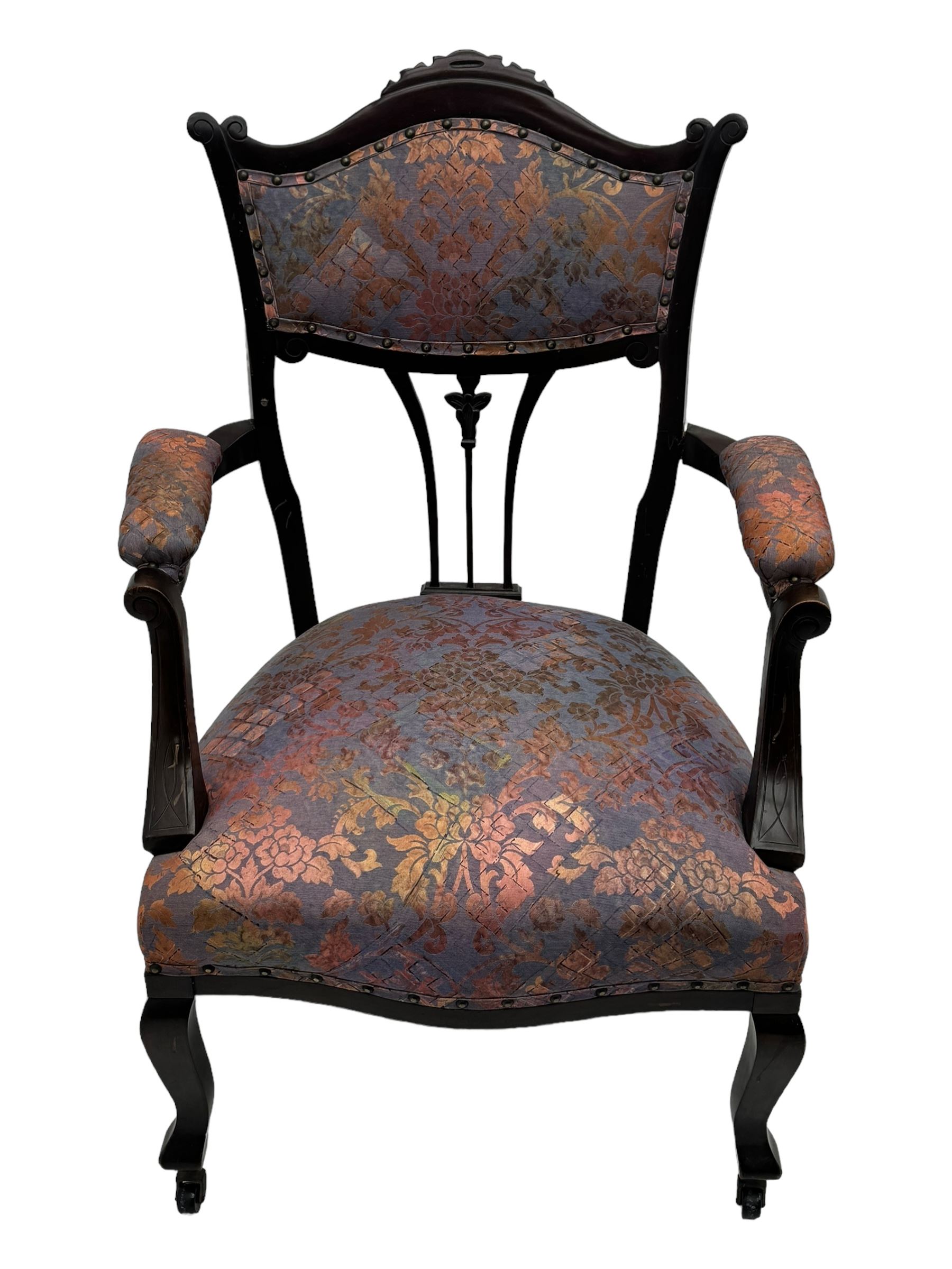 Late Victorian mahogany framed armchair, upholstered in muted purple fabric with iridescent floral pattern and nailhead trim, central foliate carved splat over padded scrolled arms, on cabriole supports fitted with castors