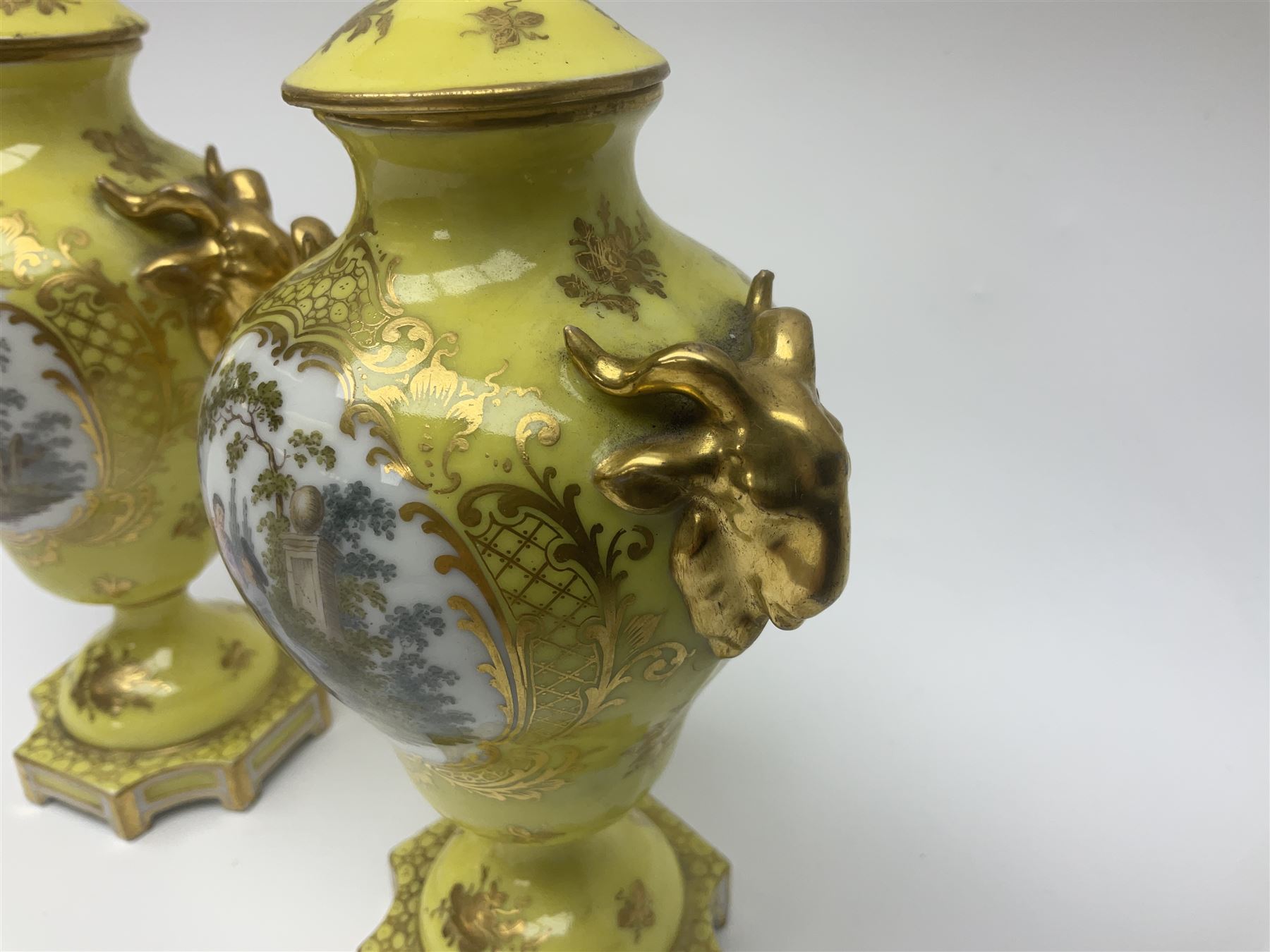 Pair of early 20th century Dresden vases with cover, of baluster form, decorated with figural panels within ornate gilt borders upon a yellow ground, with twin gilt goat mask handles, H16.5cm