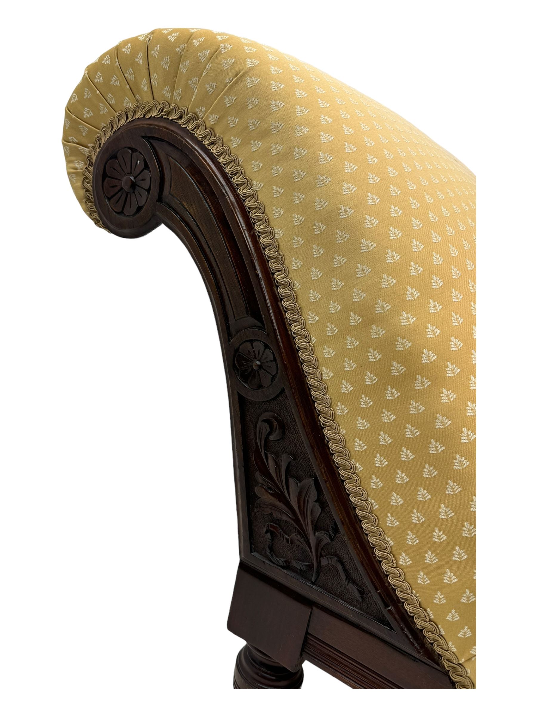 Late Victorian walnut framed chaise longue, scrolled backrest and sides upholstered in patterned yellow fabric, carved floral motifs to the backrest and sides, raised on turned supports with castors