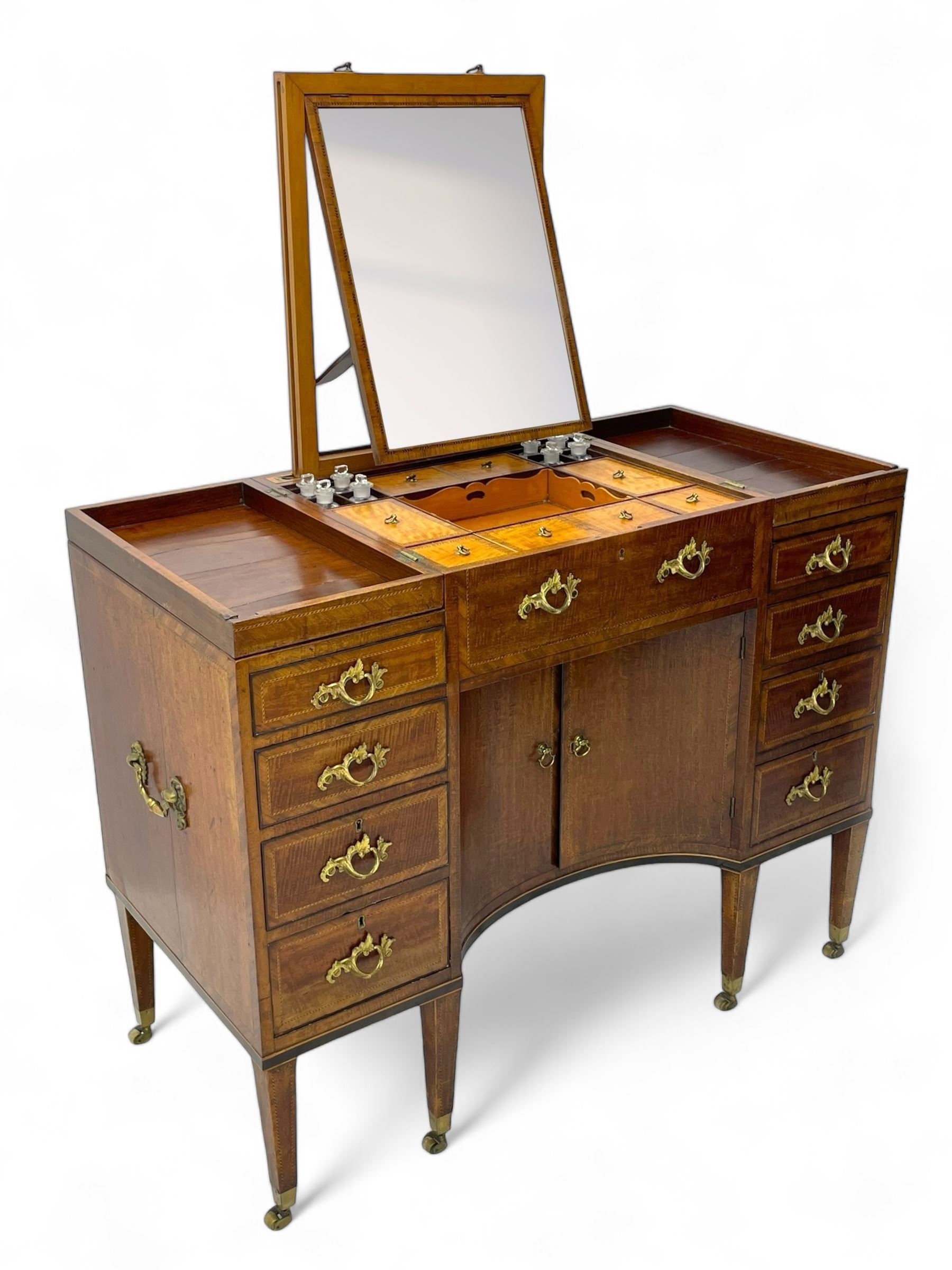 Late 19th century mahogany and satinwood dressing chest, the raised middle section hinges to reveal an interior fitted with lidded compartments, removable tray, single drawer and glass scent bottles, lift-up bevelled mirror with adjustable hinged mechanism, fitted with a further eight cock-beaded graduating drawers and curved kneehole with double cupboard, square tapering supports on brass cups and castors, inlaid throughout with checkered stringing, ornate Rococo cast gilt metal handles