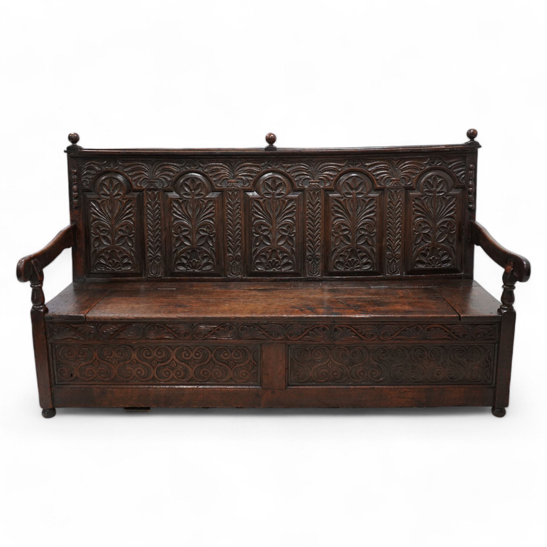 18th century oak settle or bench, moulded cresting rail with globular finials over five panel back, each stepped arch panel carved with stylised leaf decoration, hinged box seat over S-scroll foliate frieze and two panels carved with scrolling pattern, panelled sides, on stile supports terminating to turned feet 