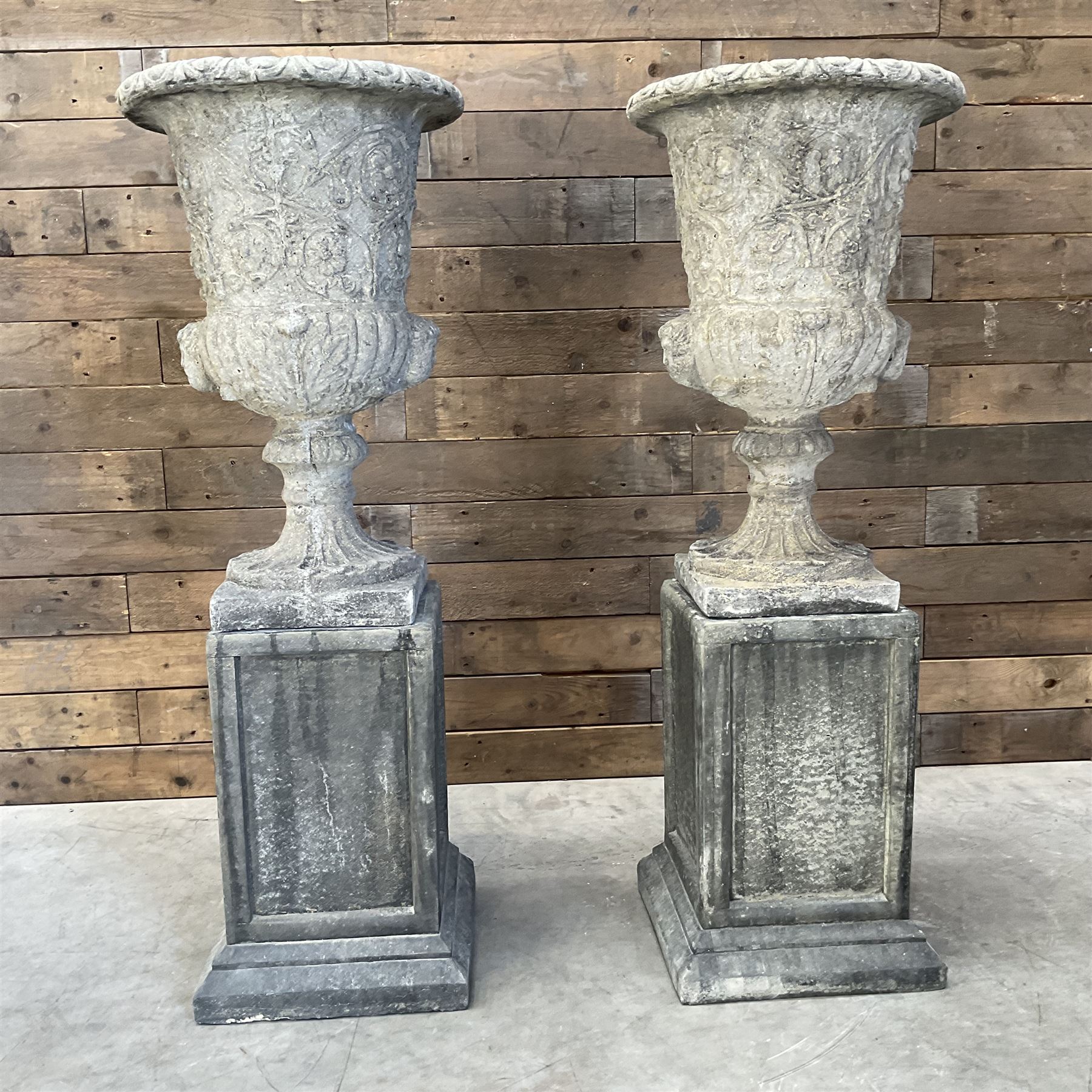 Pair of Georgian design cast stone garden urns, egg and dart rim with raised floral motifs, pedestal base terminating on square plinth, raised on square block plinth