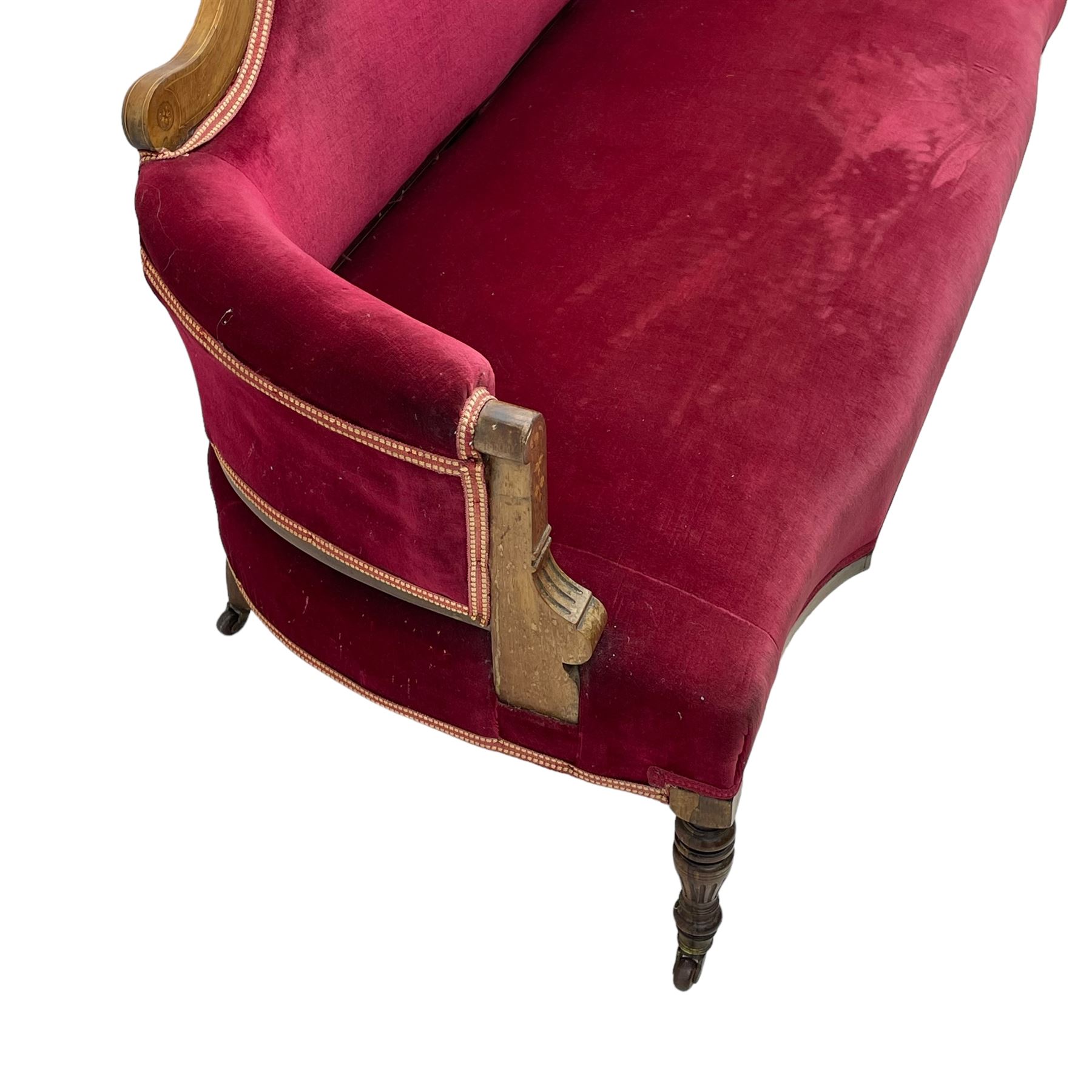 Late Victorian inlaid walnut two seat settee, the shaped cresting rail decorated with foliate inlays and boxwood stringing, upholstered in crimson velvet with sprung seat, raised on turned and fluted supports with ceramic castors