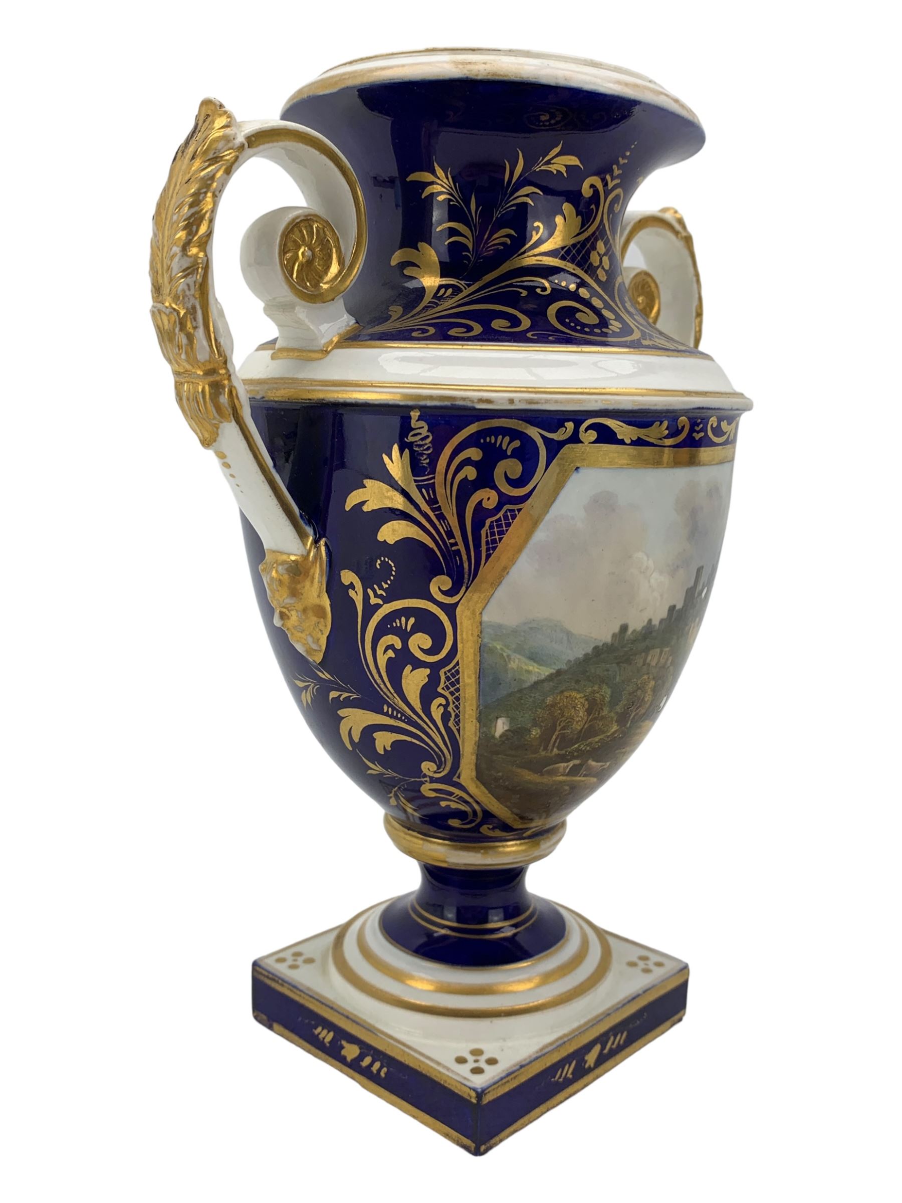 Early 19th century Derby porcelain urn, the reserve painted with a pastoral landscape and castle in the distance, with twin scroll and mask handles, upon a square pedestal base, H23cm, together with a Derby shell-shaped dish, centrally hand painted with a landscape scene titled 'In North Wales', within a cobalt blue and gilt border, L25cm (2)