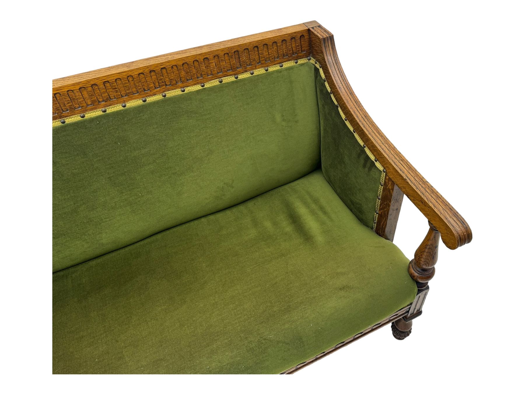 Late Victorian walnut framed sofa, rectangular backrest with carved detailing and central motif, upholstered in olive green fabric with brass studded trim and patterned front rail, scrolled fluted arms supported by turned uprights terminating in bun feet