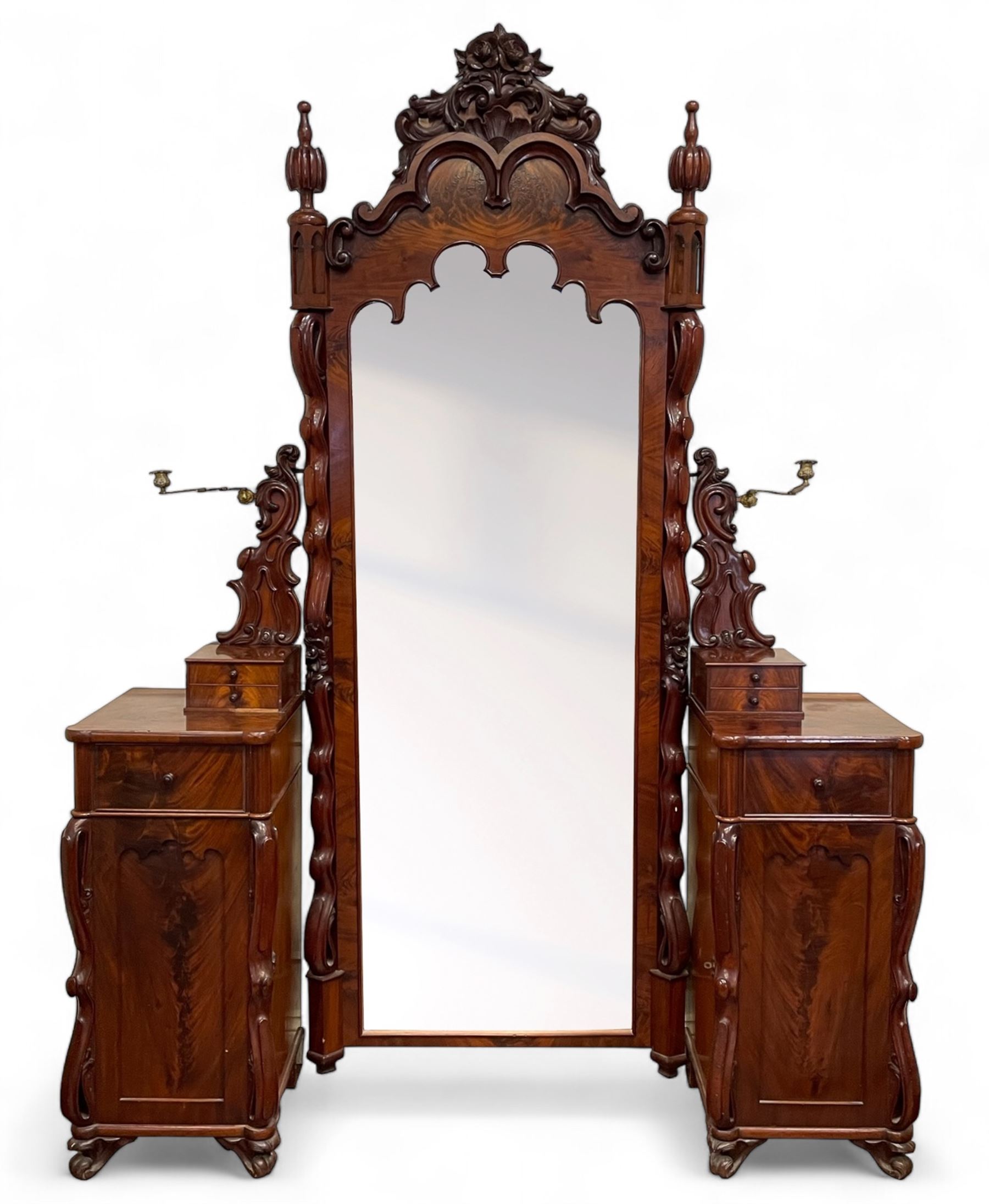 Tall Victorian figured mahogany twin pedestal dressing table, the tall mirror with flower head and scrolled leaf carved pediment, flanked by two turned and carved finials over faceted lancet arched lantern tops, plane mirror plate enclosed by shaped frame, pierced and undulating moulded uprights, on C-scroll and curled leaf carved supports fitted with cast brass hinged candle sconces, each pedestal fitted with two small trinket drawers over larger drawer and panelled cupboard, canted corners with matched upright mounts, the cupboard doors enclosing slides, on scroll carved feet 