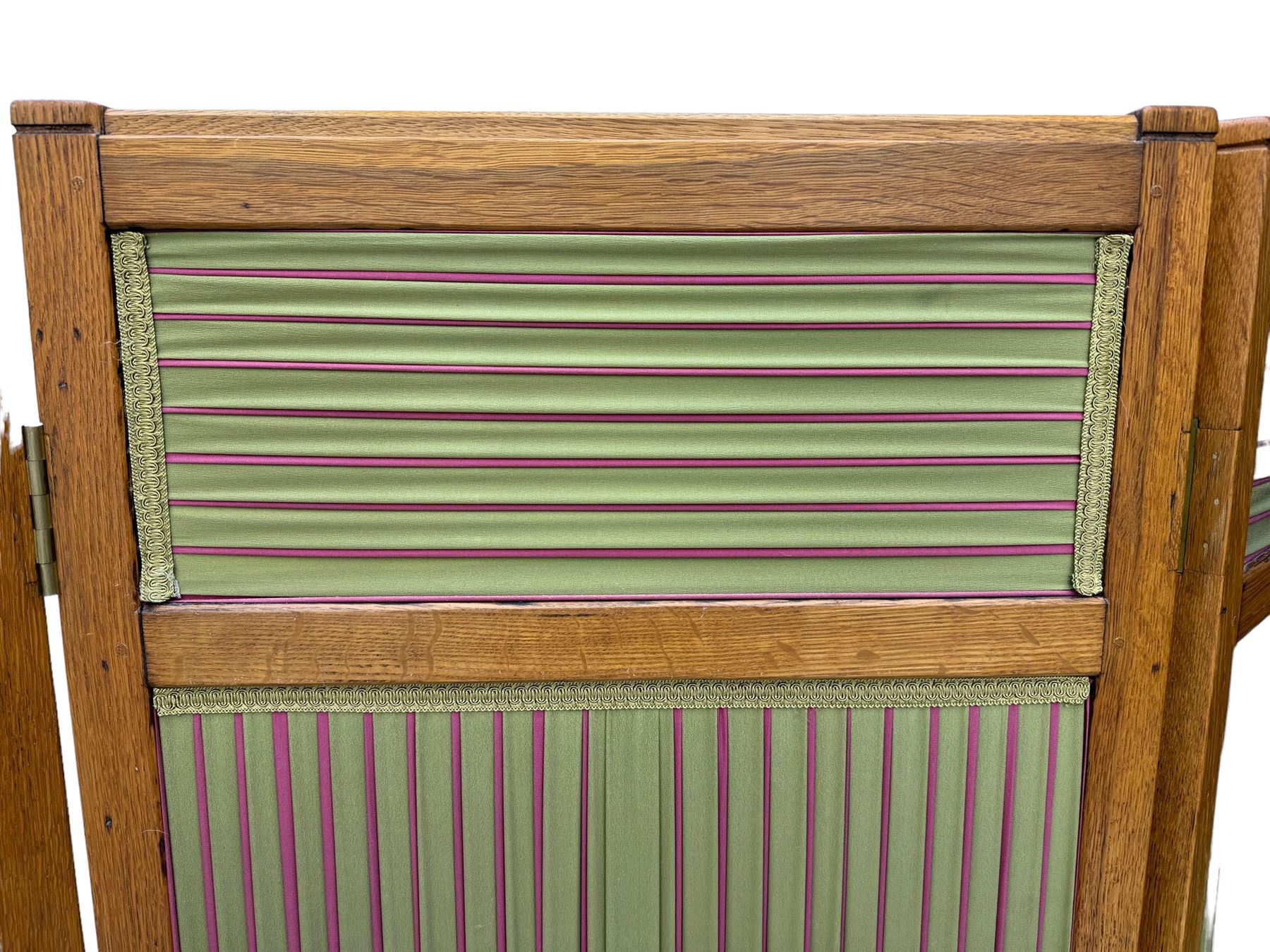 Edwardian oak and fabric folding screen, featuring three panels with oak frames, one side covered in green striped fabric with decorative trim, the reverse side in pink moiré effect fabric, connected by brass hinges