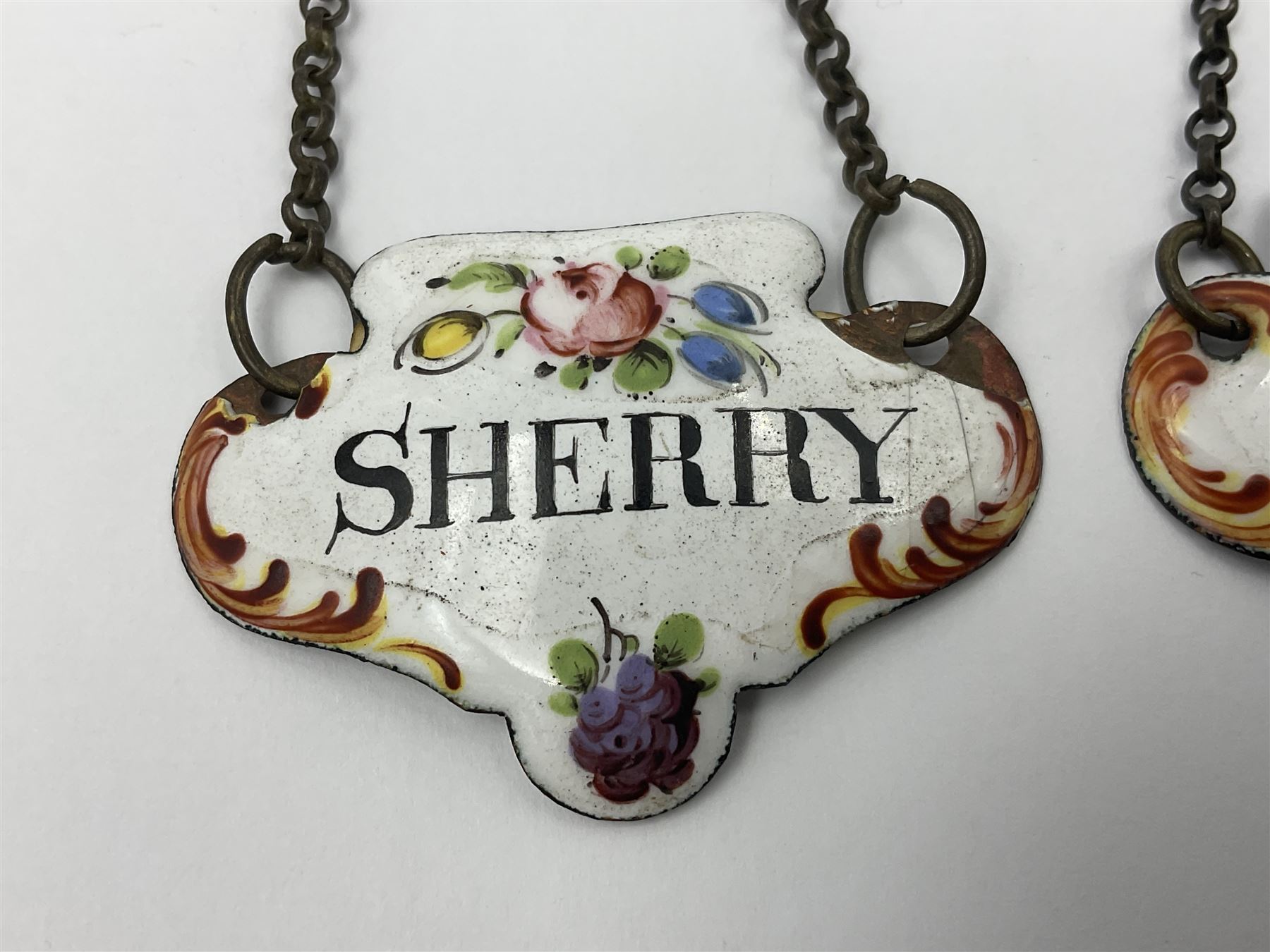 Two late 18th/early 19th century enamel wine labels, each of shaped form, titled 'CLARET', and 'SHERRY' and decorated with floral sprigs and scroll detail upon a white ground, each with suspension chain, each approximately H3.5cm W5cm