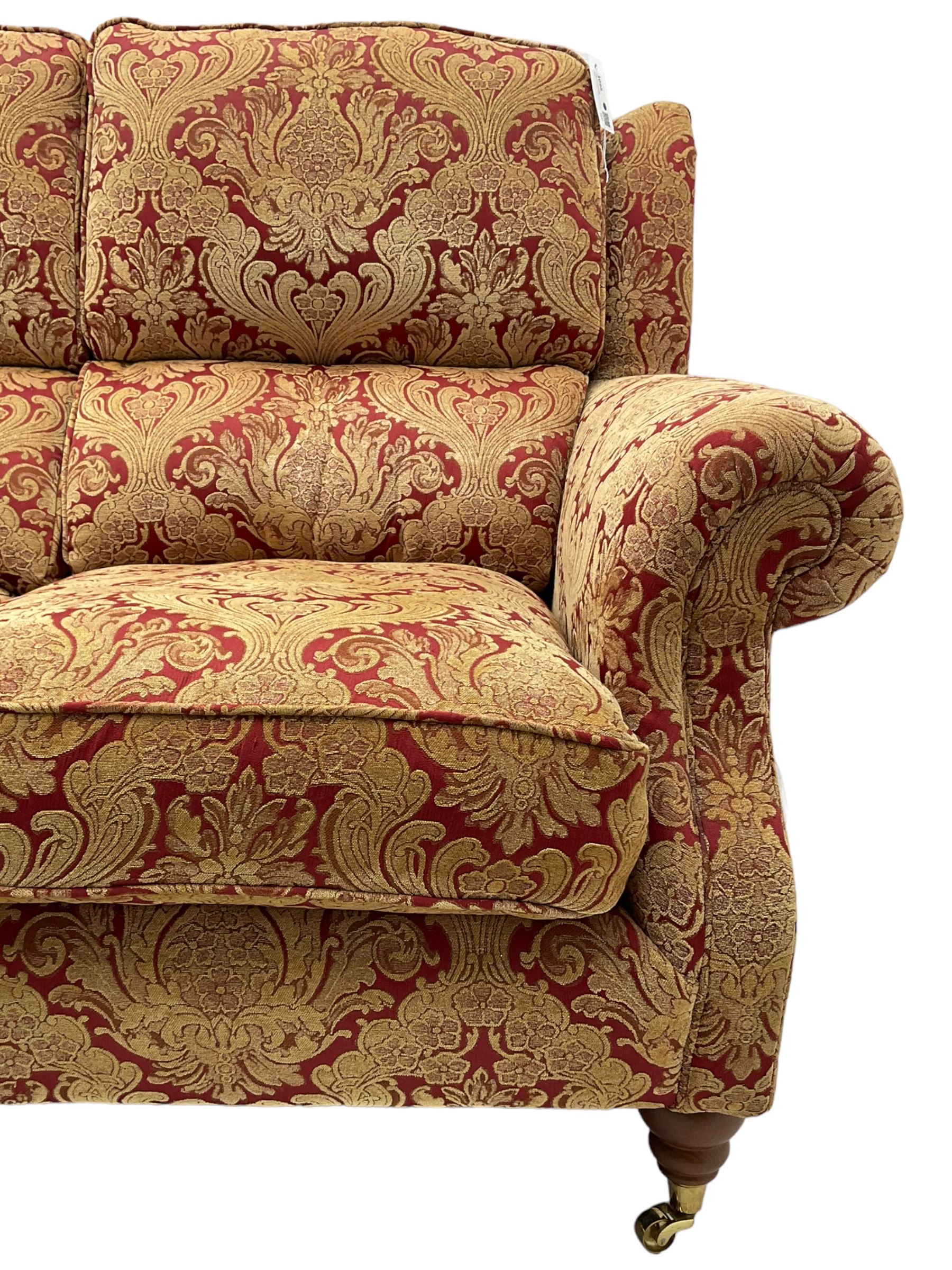 Parker Knoll - three-seat 'Oakham' hardwood-framed sofa, upholstered in red and gold floral pattern fabric, on turned feet with brass cups and castors 