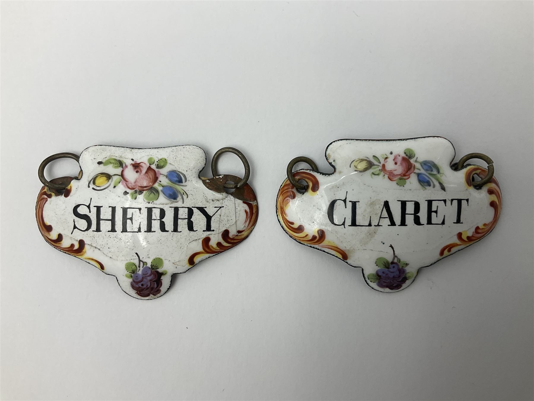 Two late 18th/early 19th century enamel wine labels, each of shaped form, titled 'CLARET', and 'SHERRY' and decorated with floral sprigs and scroll detail upon a white ground, each with suspension chain, each approximately H3.5cm W5cm