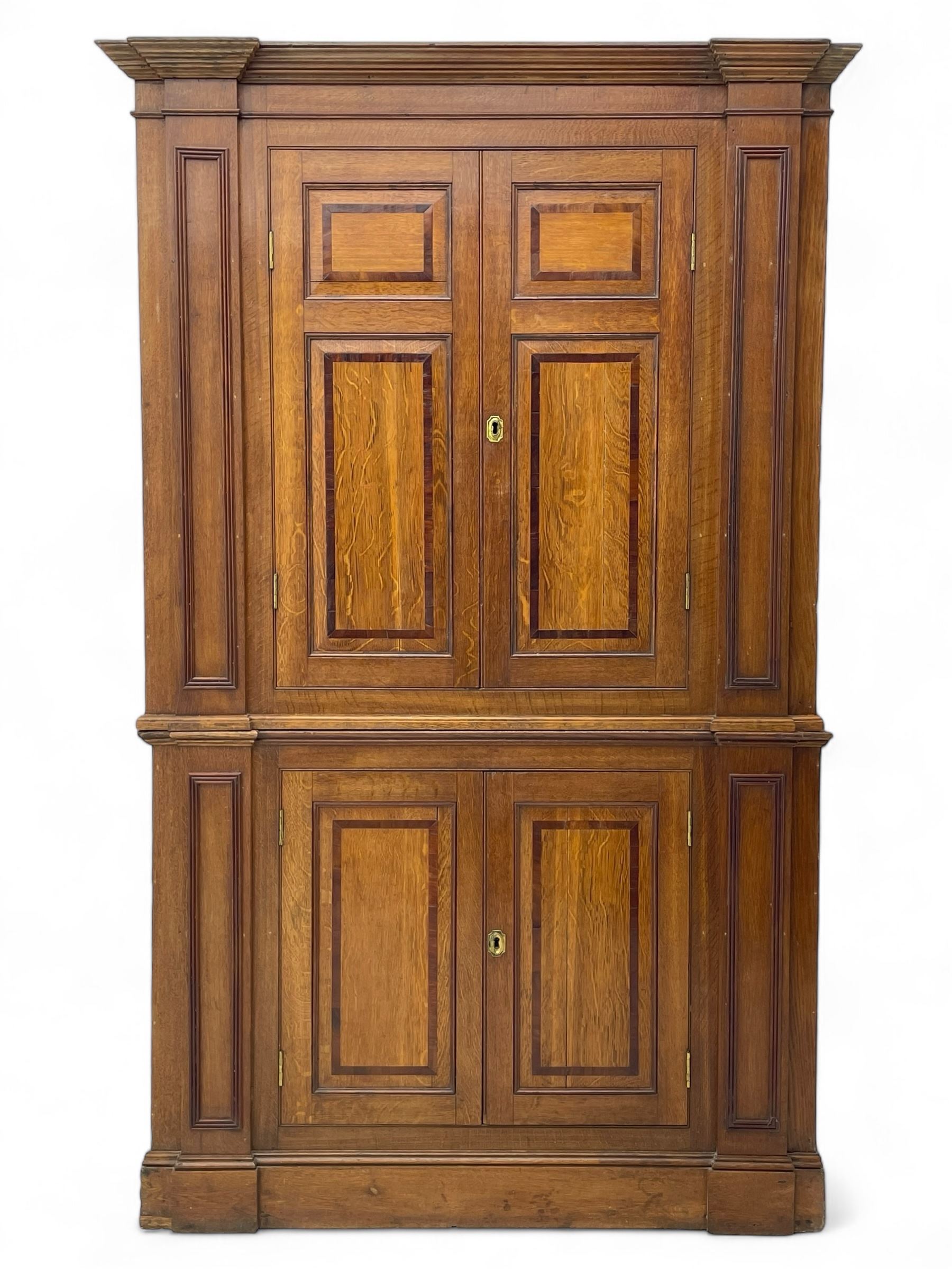 George III and later oak and mahogany banded corner cupboard, moulded cornice over four panelled doors, the door panels inlaid with mahogany bands, projecting square pilasters throughout with applied reeded mouldings, the painted interior fitted with shaped shelves, on moulded plinth base 