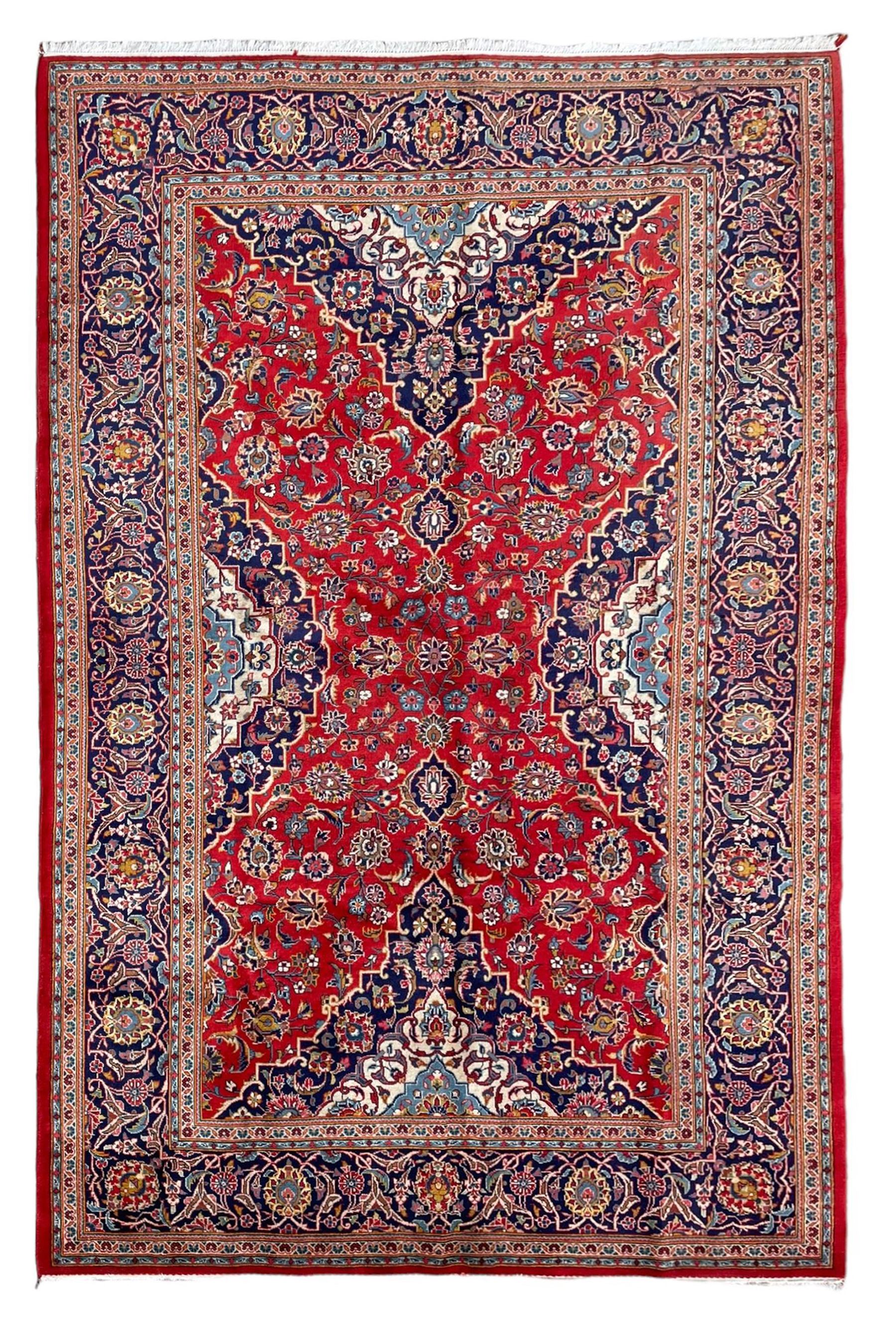 Persian Kashan crimson ground rug, overall arabesque design, the field decorated with swirled leafy branches and palmettes, indigo ground border decorated with repeating pattern, within guard stripes 