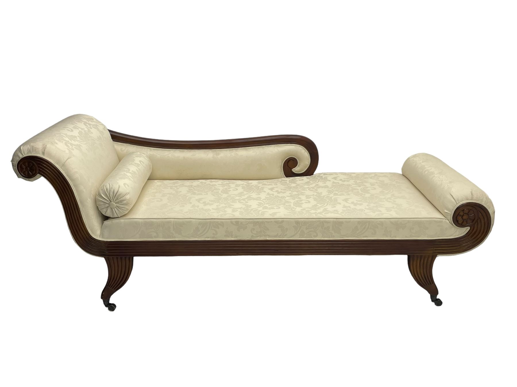 Regency design mahogany chaise longue, scrolled arms with carved rosette details, upholstered in cream damask fabric with bolster cushion, reeded frame supported by turned legs on brass castors