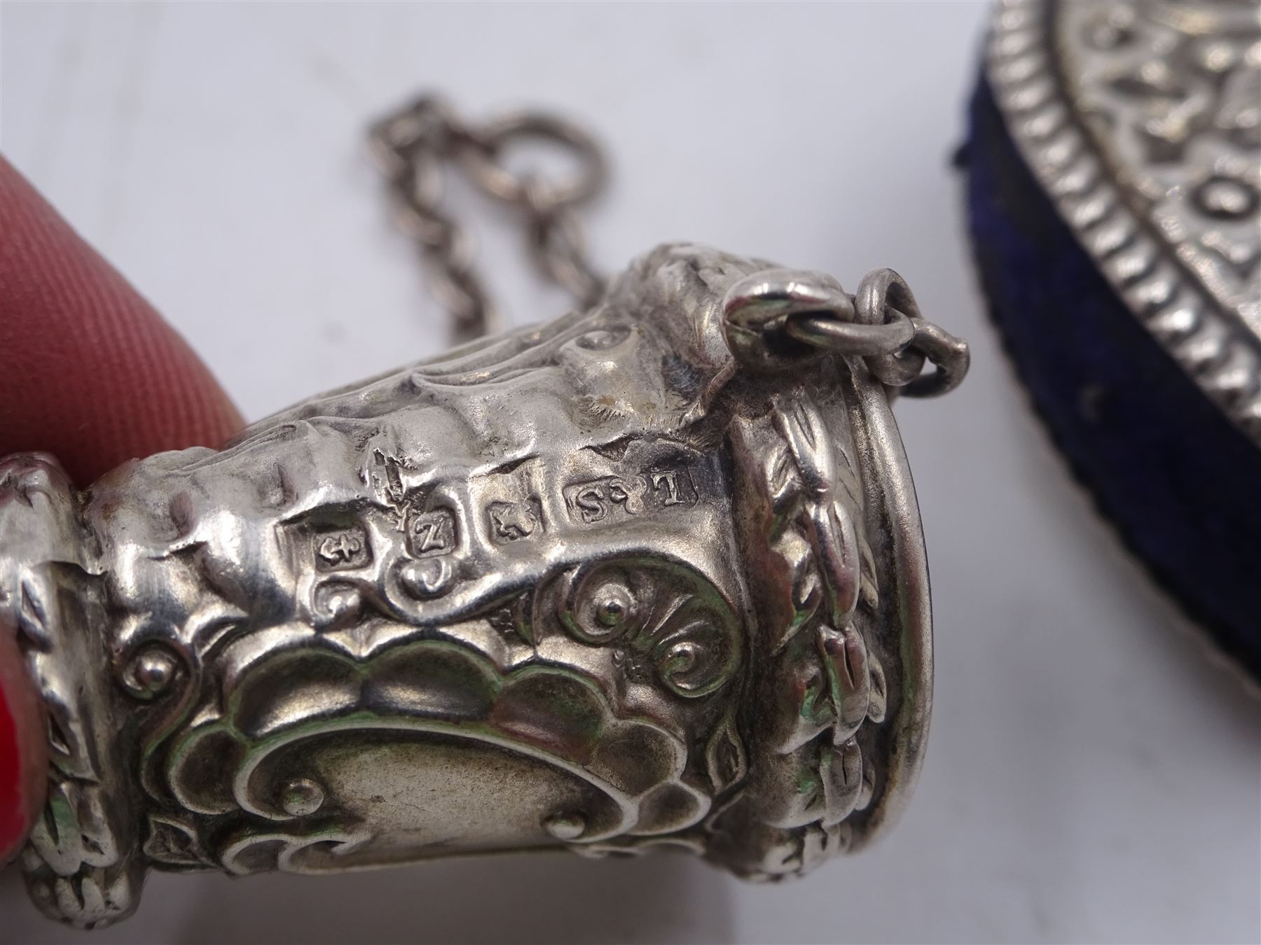 Edwardian silver pin cushion, in the form of a heart, with embossed floral decoration, with blue pin cushion between two heart shaped silver covers, with chatelain hook, hallmarked Henry Matthews, Birmingham 1902, together with a late Victorian silver thimble holder, embossed with scrolling decoration and with velvet interior, with chatelain chain, hallmarked Levi & Salaman, Birmingham 1899, pin cushion H4.5cm