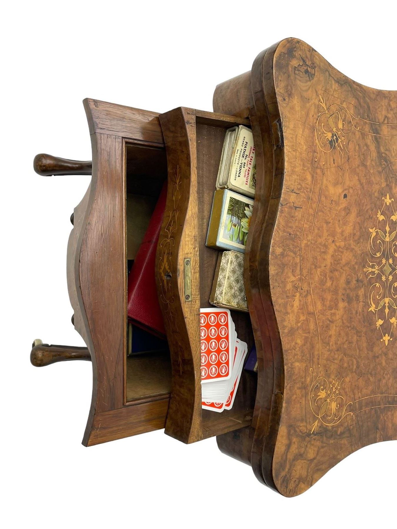 Victorian inlaid figured walnut games table, the serpentine fold-over top inlaid with scrolling foliate decoration, the top opens to reveal cribbage, backgammon and chess boards, single drawer over sliding storage well, on twin pillar supports united by double turned stretchers, on foliage carved out-splayed supports with brass and ceramic castors 