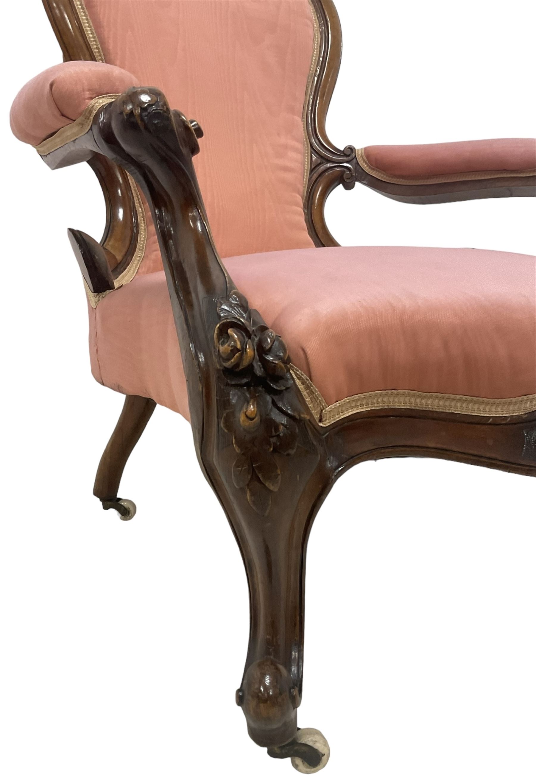 Victorian mahogany framed open armchair, spoon-back and sprung seat upholstered in pink fabric, scrolled arm terminals over serpentine fronted seat decorated with moulded carved floral motifs, cabriole supports with ceramic castors